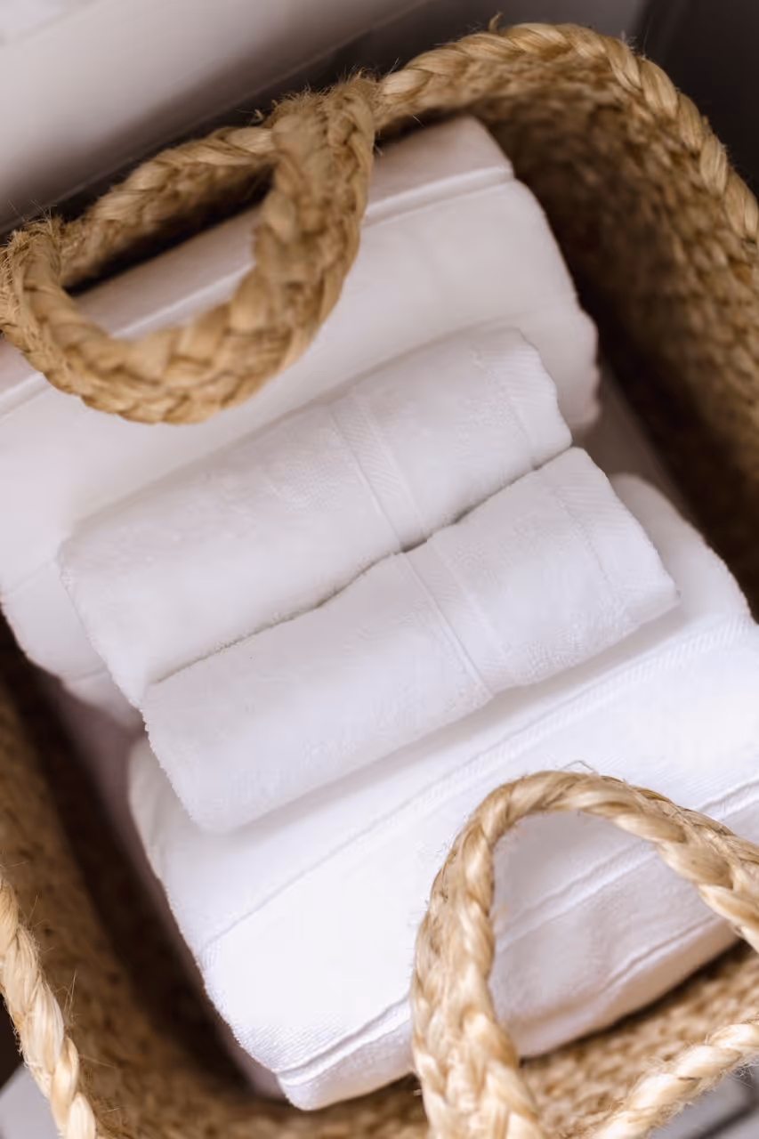 Two rolled white towels placed on top of folded white towels inside a woven basket with rope handles.