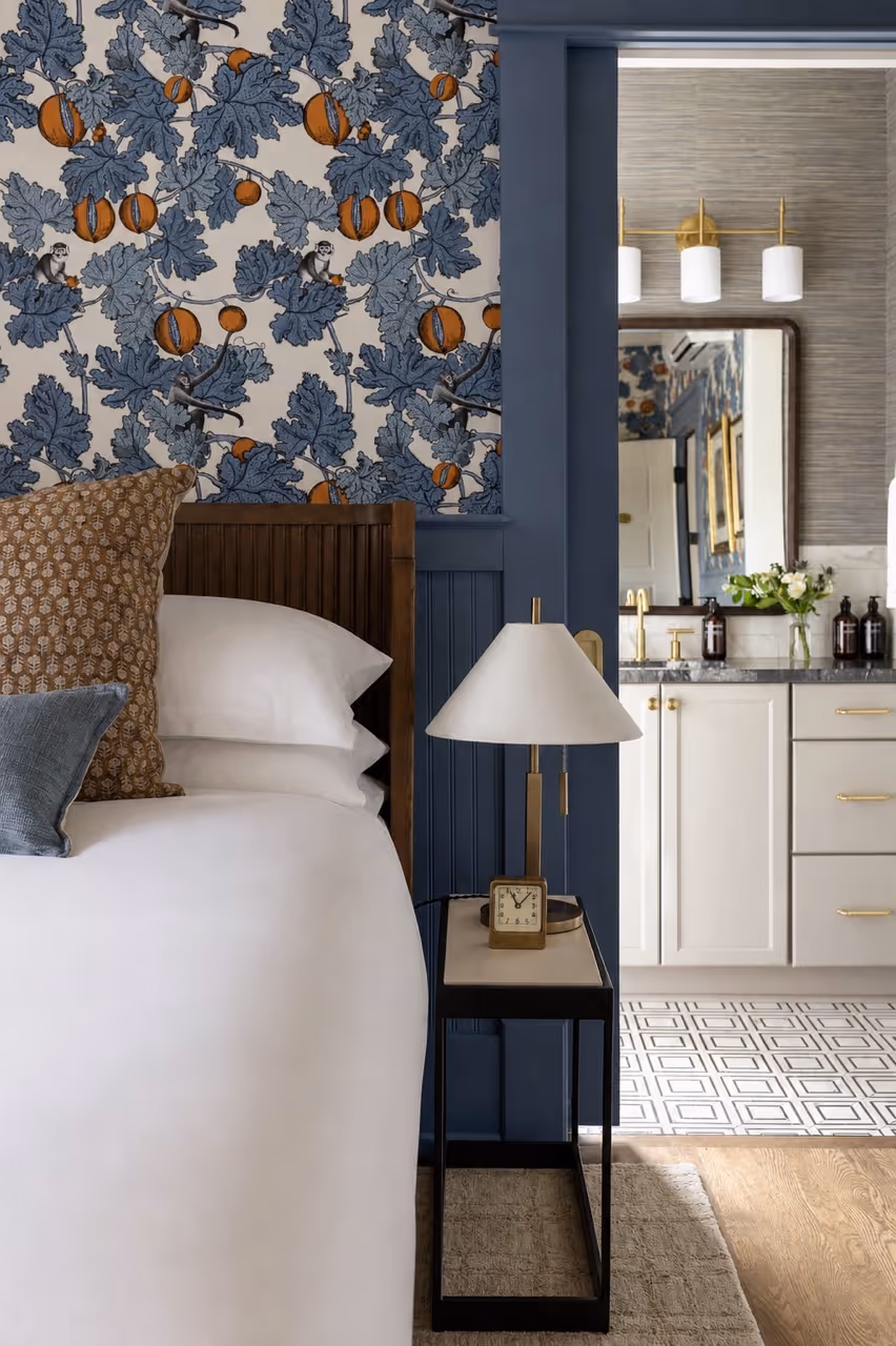 Bedside table with a lamp and clock next to a bed with patterned pillows, blue and orange botanical wallpaper behind.