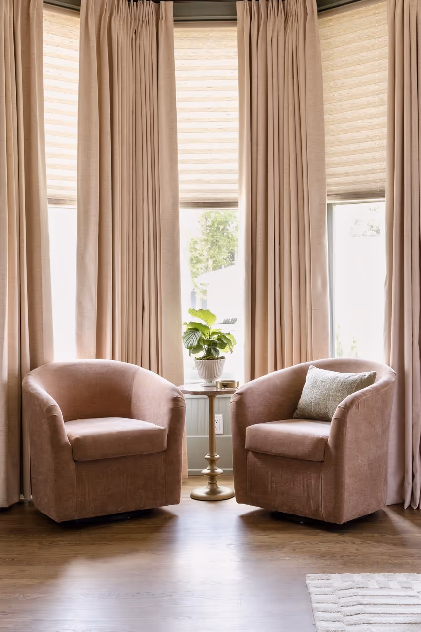 Two blush pink armchairs flanking a small round table with a potted plant, set in front of tall beige curtains.