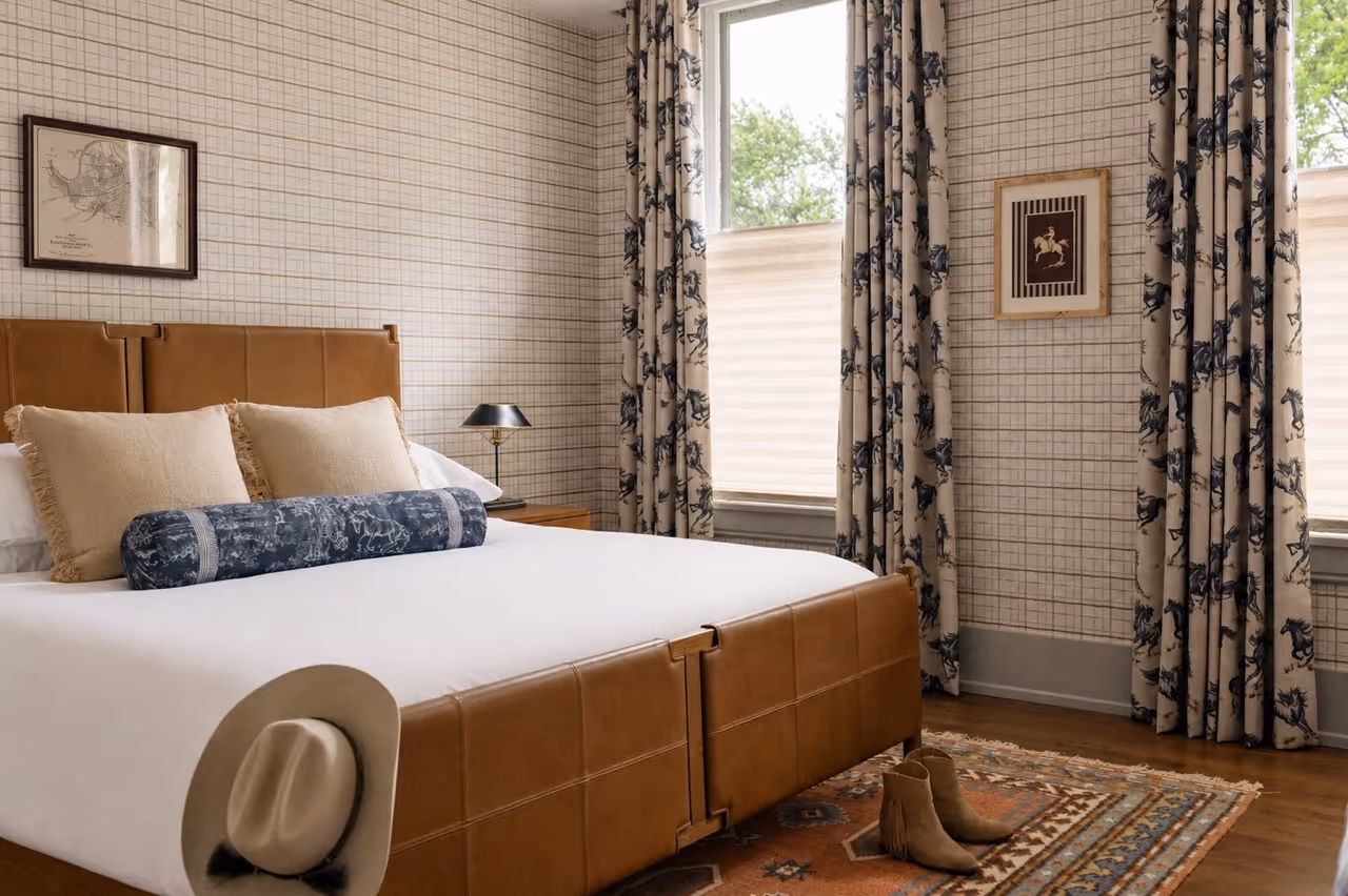 Bedroom with a brown leather bed, beige pillows, patterned curtains, a hat on the bed corner, and boots on a patterned rug.