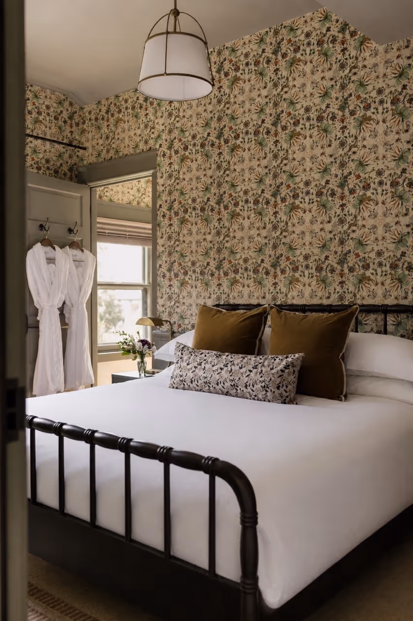 Bedroom with black metal bed frame, white bedding, brown pillows, floral wallpaper, and two white bathrobes hanging on door.