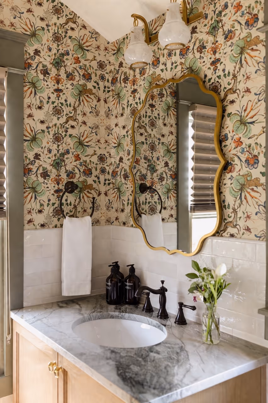Bathroom sink with marble countertop, ornate gold-framed mirror, floral wallpaper, and two black soap dispensers.