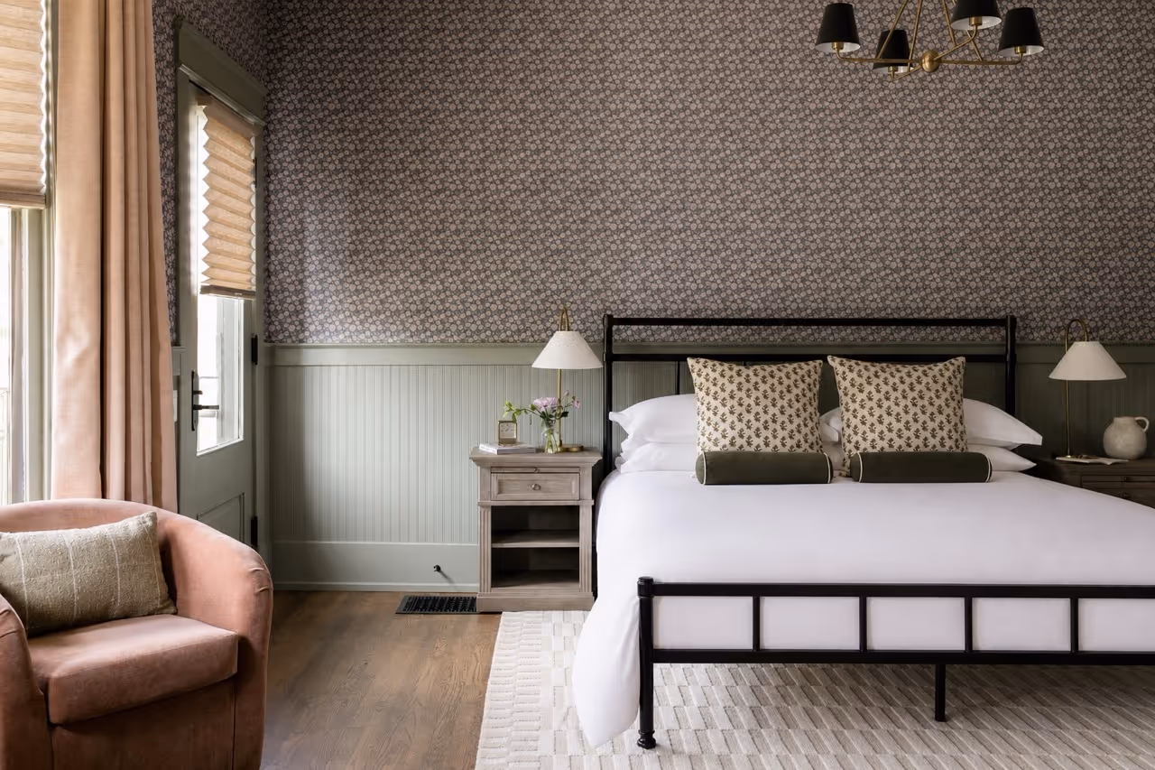 Bedroom with a black metal bed frame, white bedding, patterned pillows, floral wallpaper, wood floors, and a pink armchair.