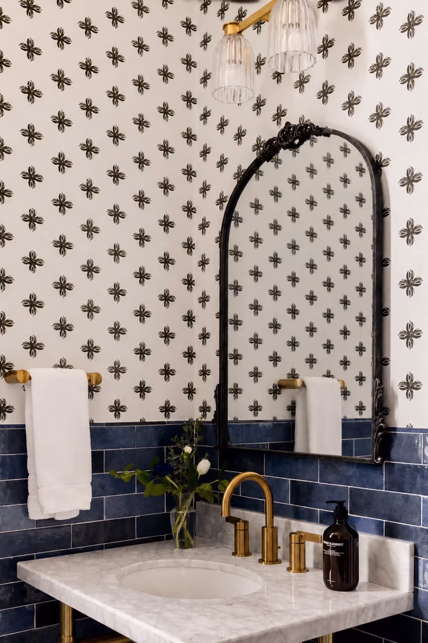 Bathroom vanity with brass faucet, oval black-framed mirror, white countertop, blue tile backsplash, and patterned wallpaper.
