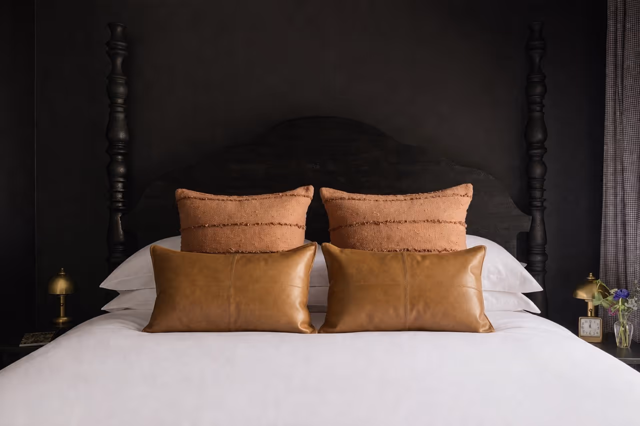 Bed with dark wooden headboard, white bedding, two brown leather pillows, and two textured brown throw pillows.