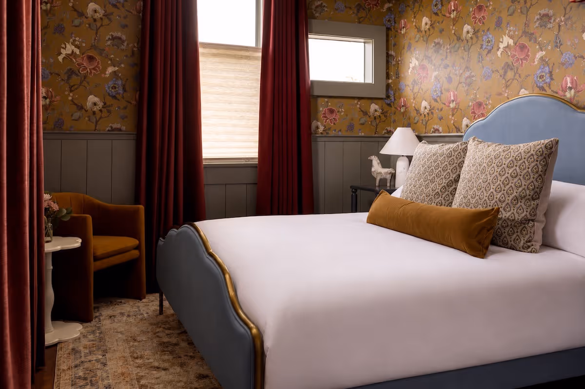 Bedroom with blue upholstered bed, patterned pillows, mustard bolster, floral wallpaper, and red curtains.
