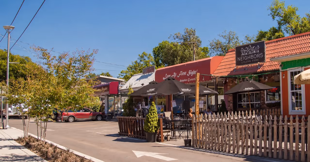 Outdoor dining area with black umbrellas, wooden fence, and small shops under clear blue sky.