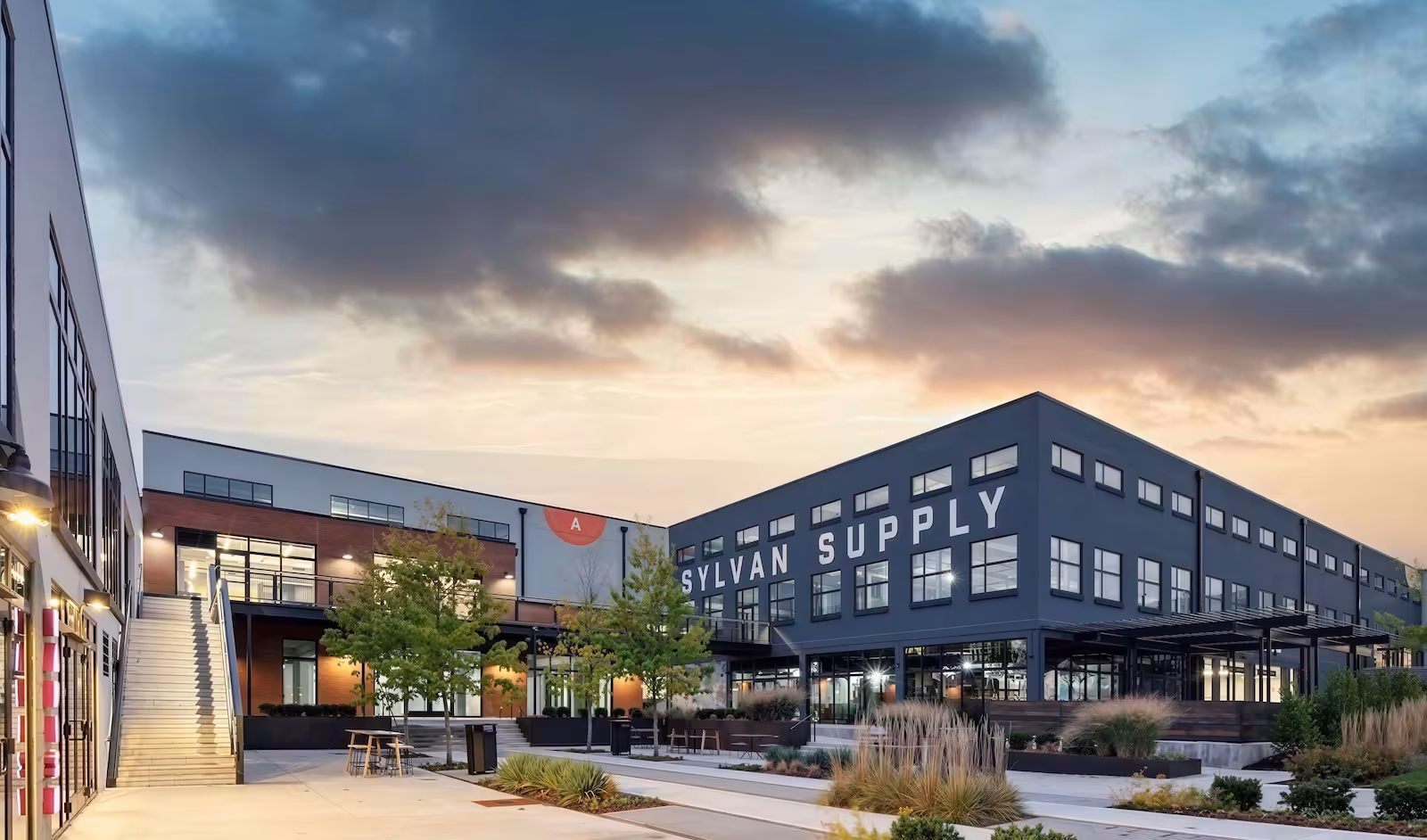 Modern commercial buildings at dusk with Sylvan Supply sign and landscaped courtyard with trees and seating.