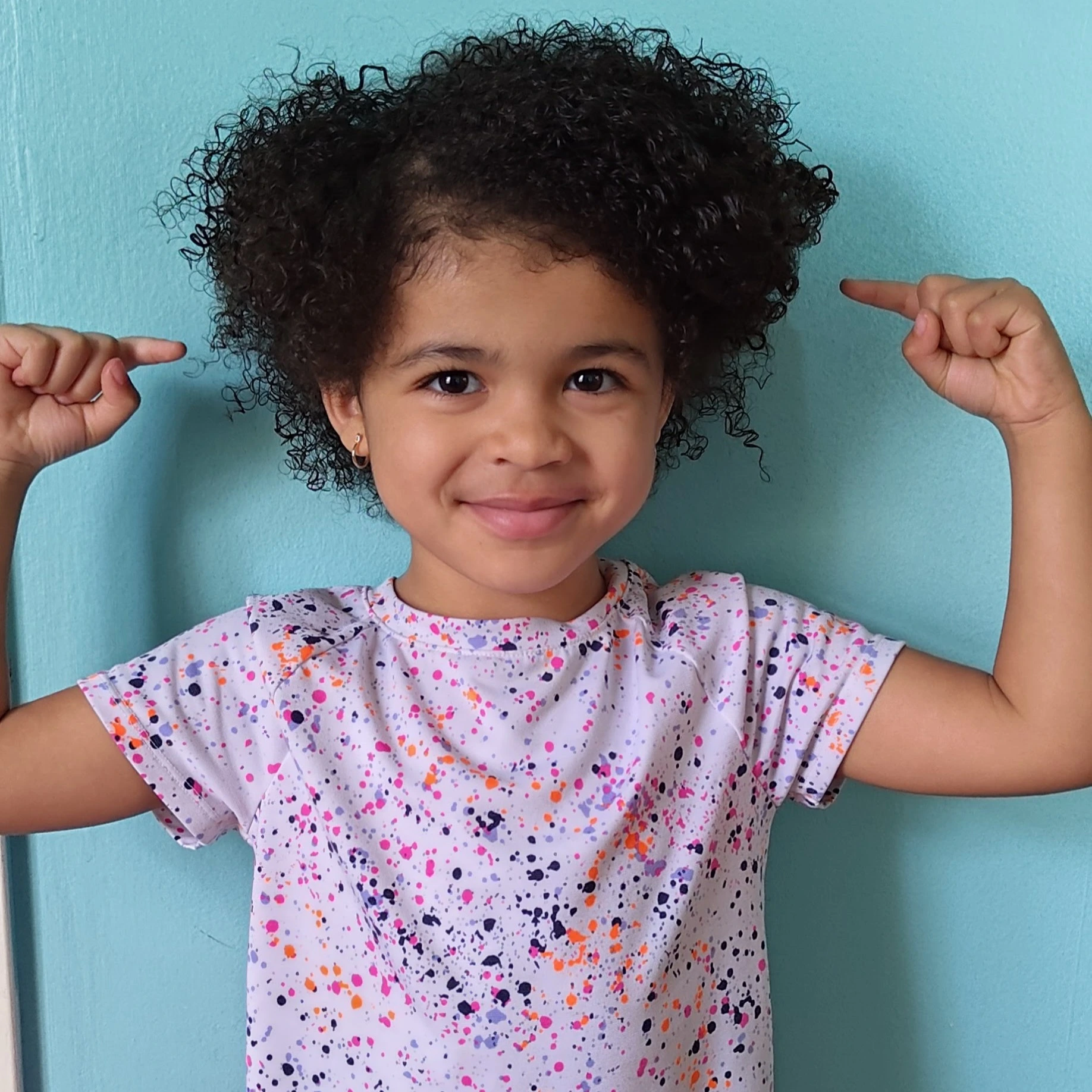 Smiling girl with curly hair wearing a colorful splatter-patterned shirt flexing her arms in front of a blue wall.