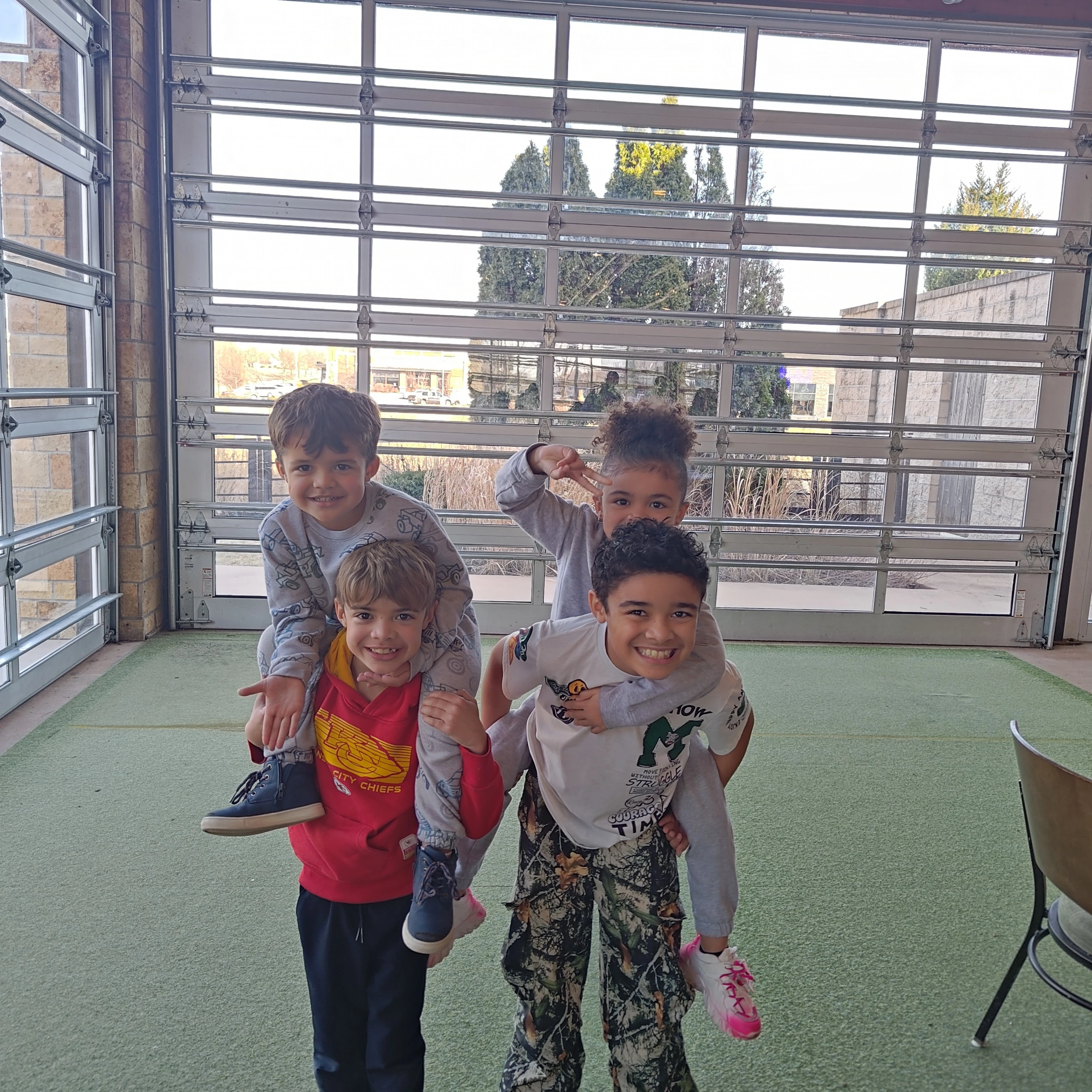 Two boys giving piggyback rides to two girls inside a garage with large windows and green carpet.
