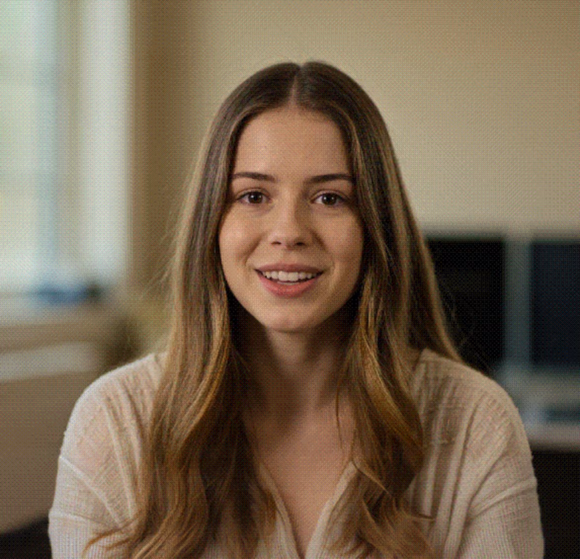 Smiling young woman with long brown hair and light sweater sitting indoors.