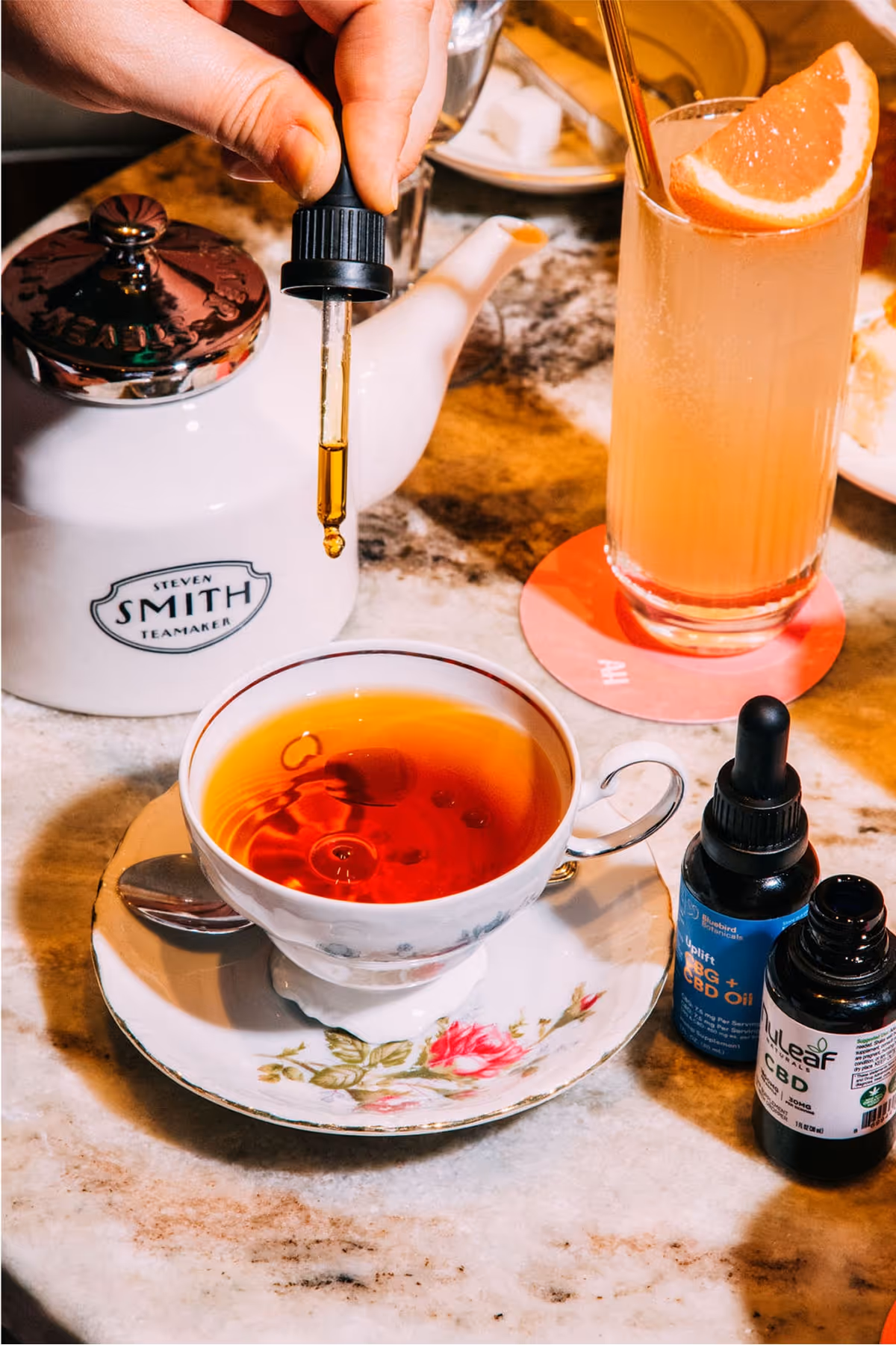 A hand holds a dropper above a teacup, adding drops to tea. A teapot, a glass of grapefruit drink, and CBD oil bottles sit on a marble table. The teacup rests on a saucer with a floral design.