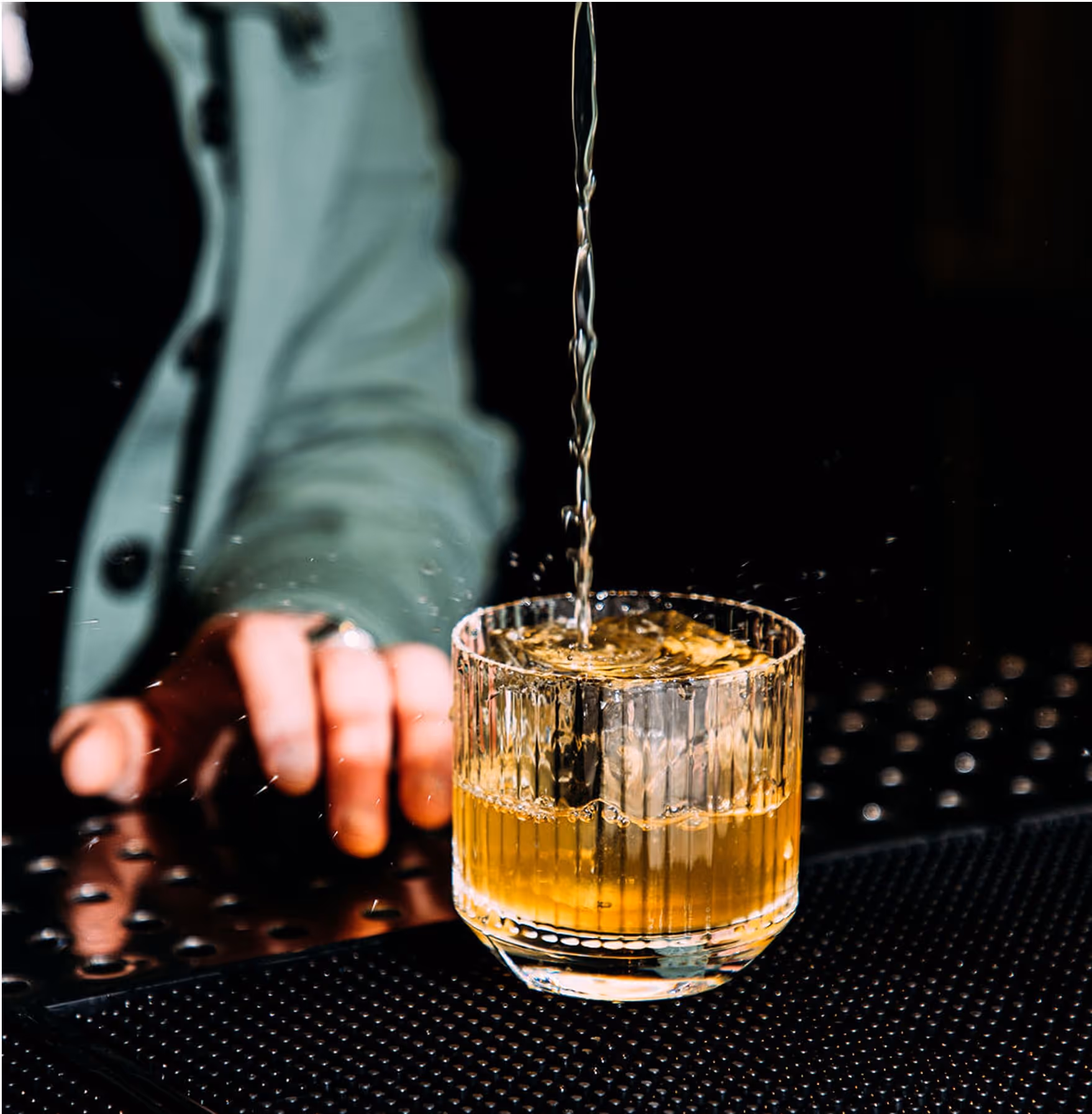 A glass of amber-colored liquid with ice is being filled, while a person in a green jacket stands in the background with their hand resting on the bar.