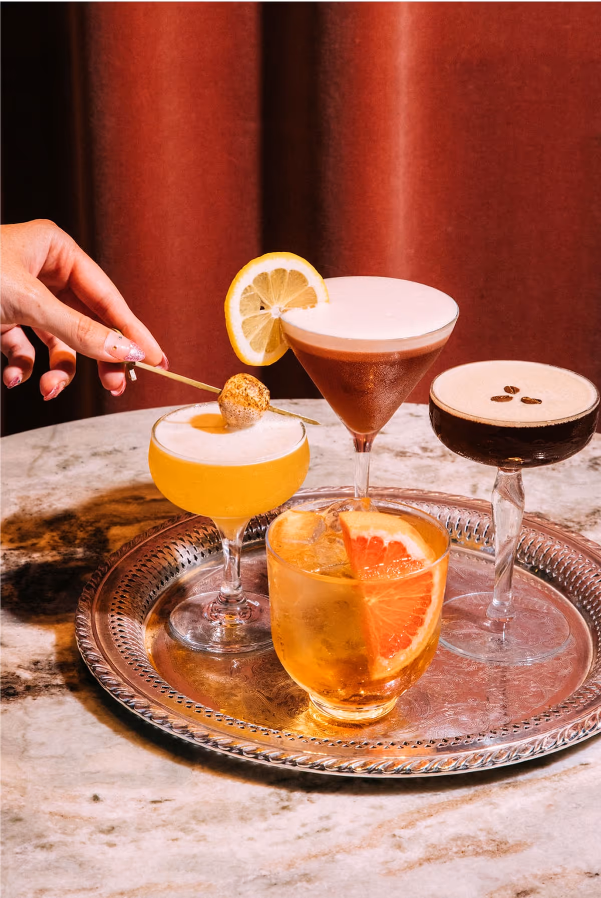 A hand garnishes a yellow cocktail with a lemon slice, among four colorful cocktails on a silver tray, including drinks with lemon, grapefruit, and coffee bean garnishes, set on a marble table with a red curtain in the background.