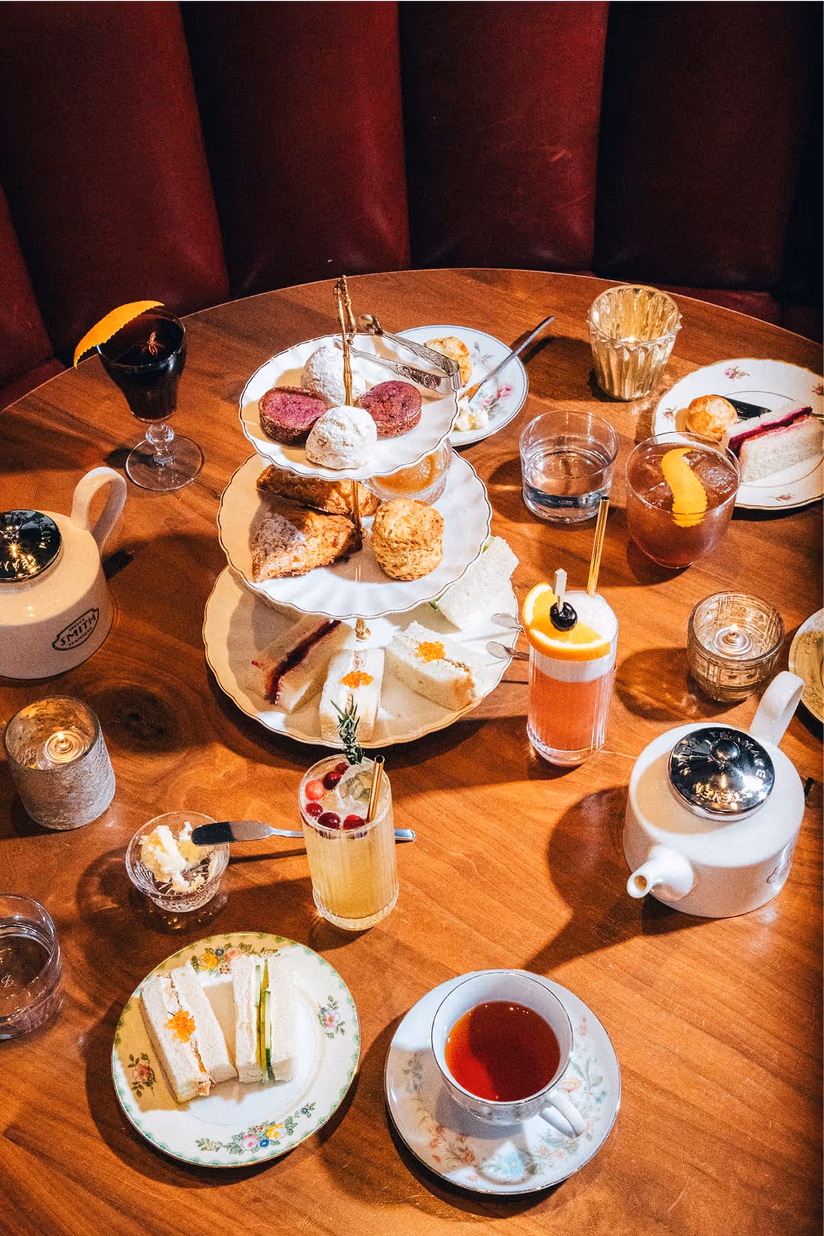 A wooden table set for afternoon tea with a three-tiered stand of pastries, scones, and desserts, teapots, cups of tea, assorted cocktails, finger sandwiches, and small plates on floral china.