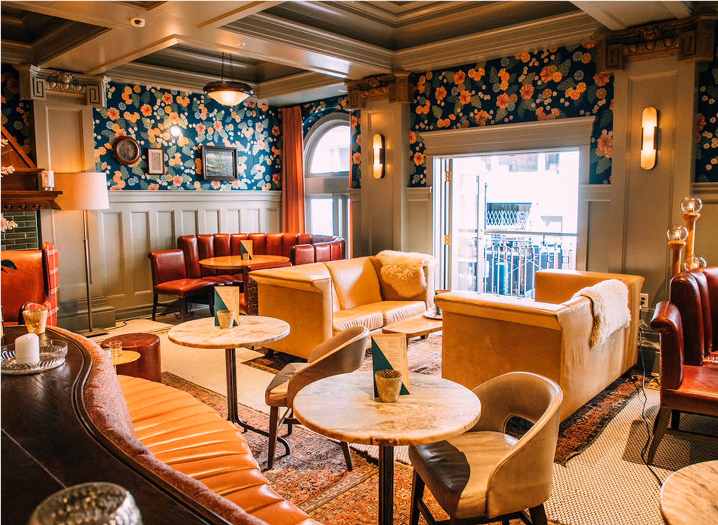 A cozy lounge with floral wallpaper, vintage furniture, tan and red leather seating, round marble tables, and large windows letting in natural light. The space has a warm, inviting ambiance and decorative accents.