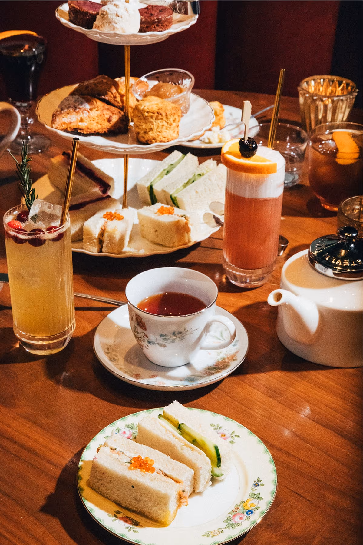 A table set for afternoon tea with a tiered tray of assorted pastries and sandwiches, a teapot, a floral teacup, two cocktails, and a small plate with finger sandwiches and cucumber slices.