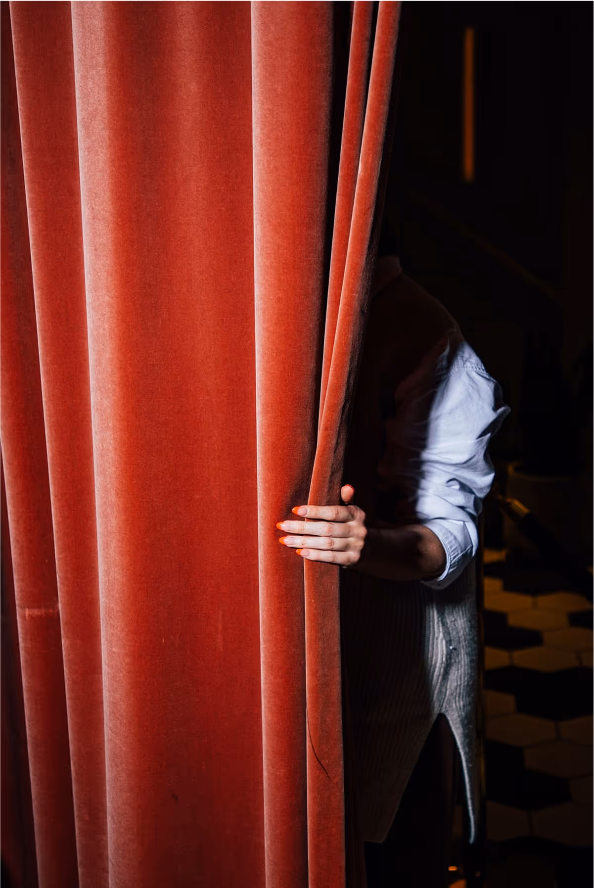 A person in a white shirt partially hides behind a heavy red velvet curtain, with their hand gripping the fabric. The background is dimly lit and features black and white floor tiles.