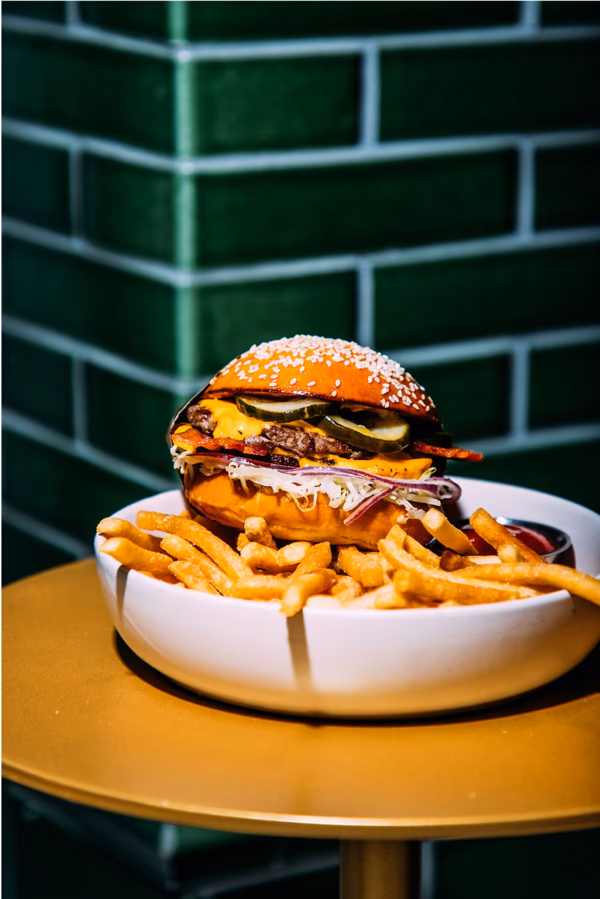 A cheeseburger with lettuce, tomato, and bacon sits on a white plate filled with French fries, set on a gold table in front of a green tiled wall.