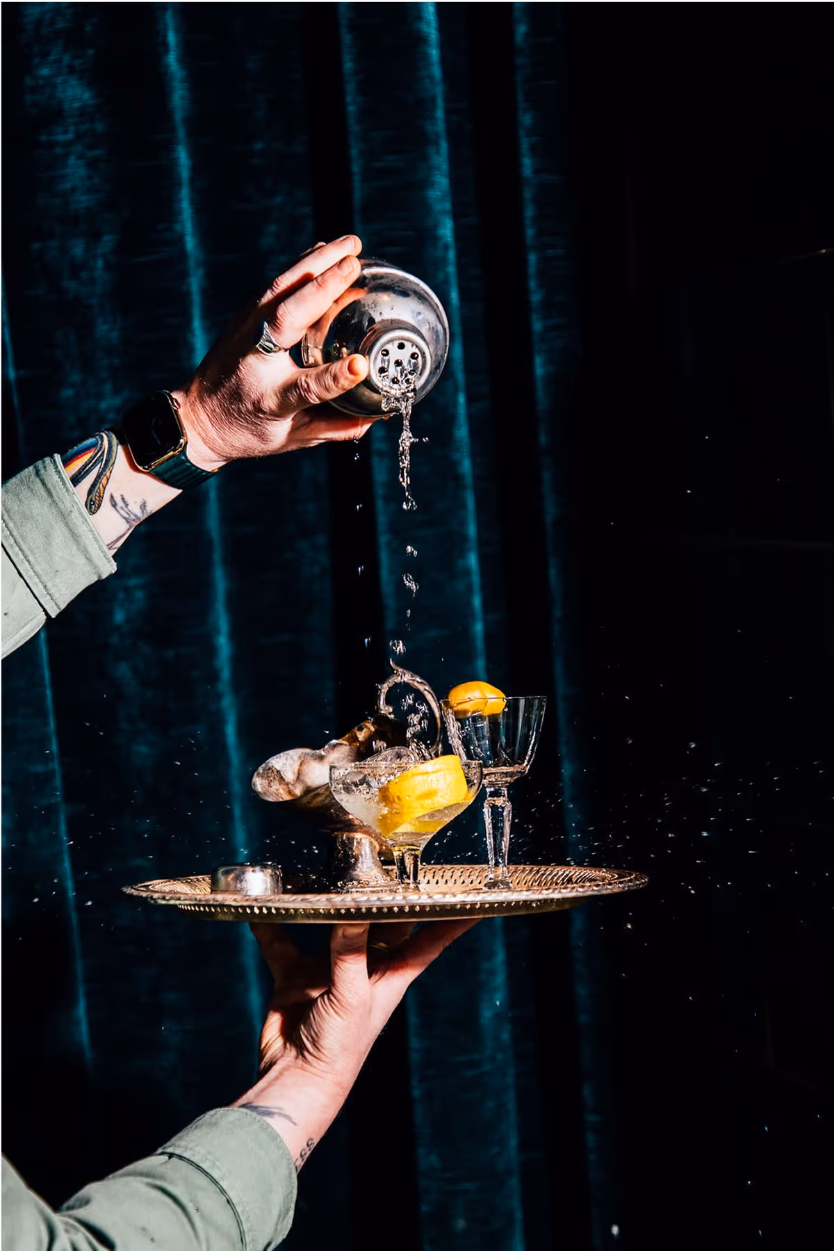 A person pours liquid from a cocktail shaker into a glass on a tray with ice and a lemon wedge, causing a splash; the scene is set against dark teal curtains.