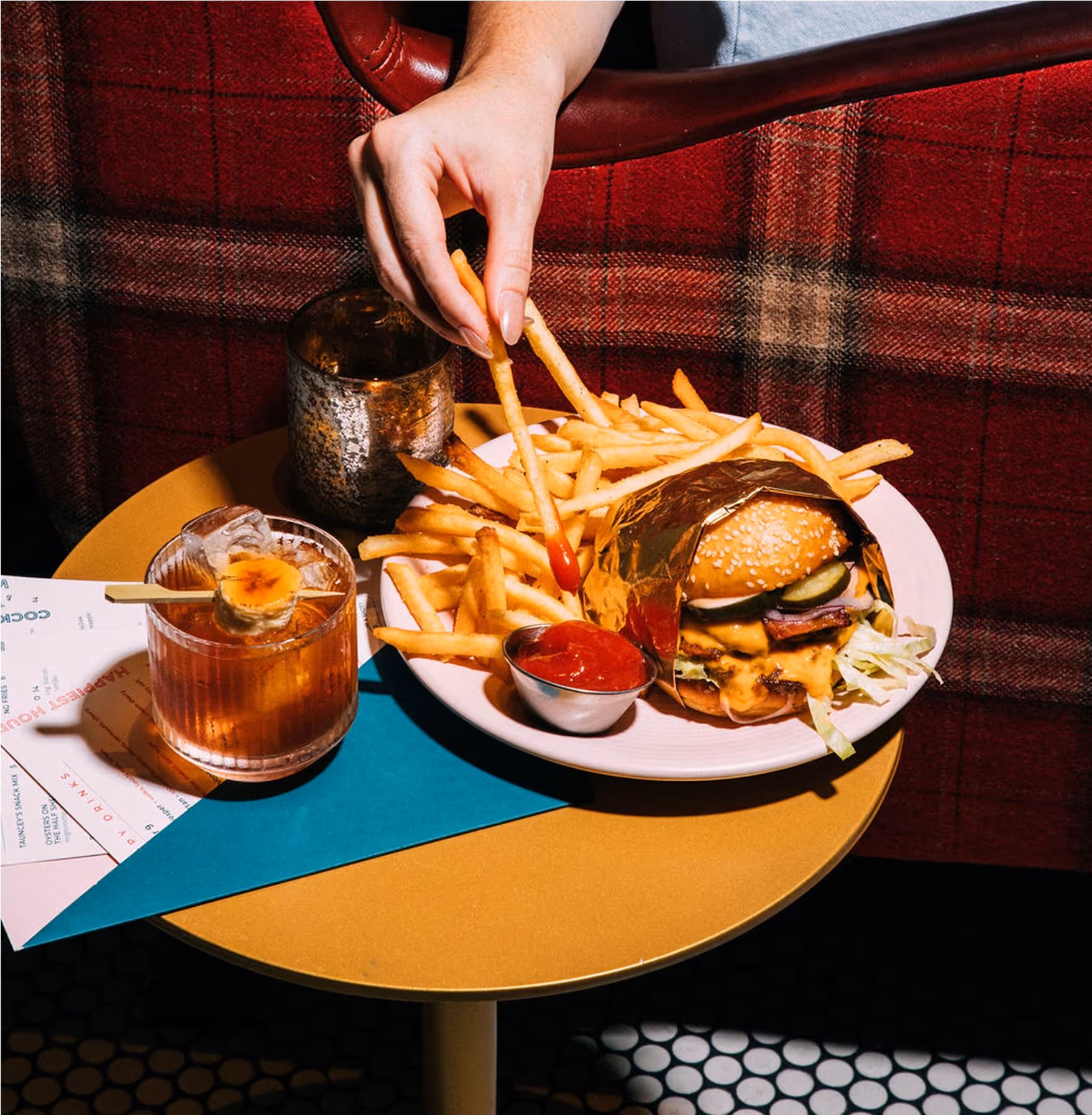 A person dips a French fry into ketchup next to a burger wrapped in foil, more fries, and a cocktail with an orange slice, all on a round yellow table in front of a red plaid background.