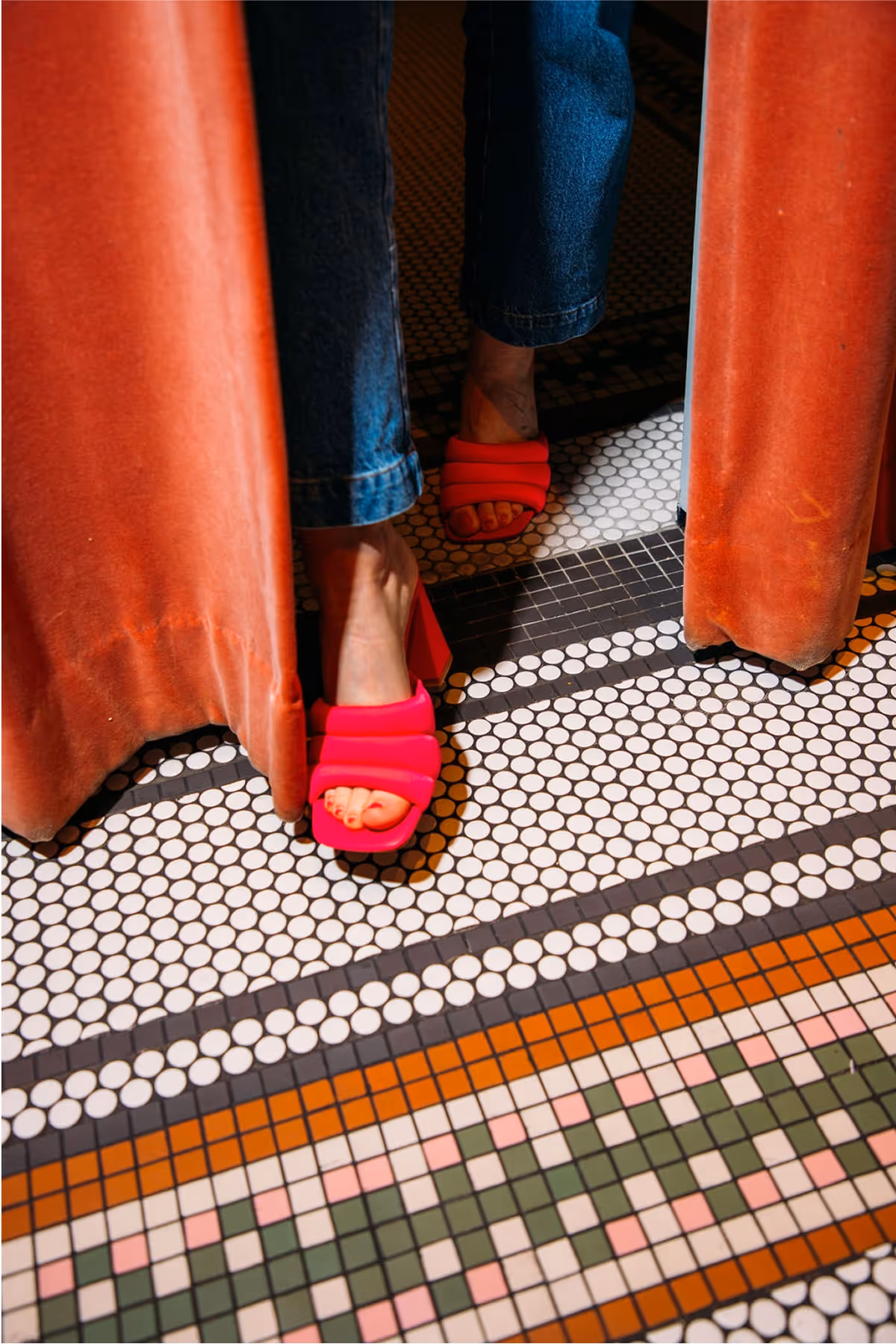 A person in blue jeans wearing bright pink heeled sandals stands behind partially opened orange curtains on a patterned tile floor with white, brown, and green tiles.