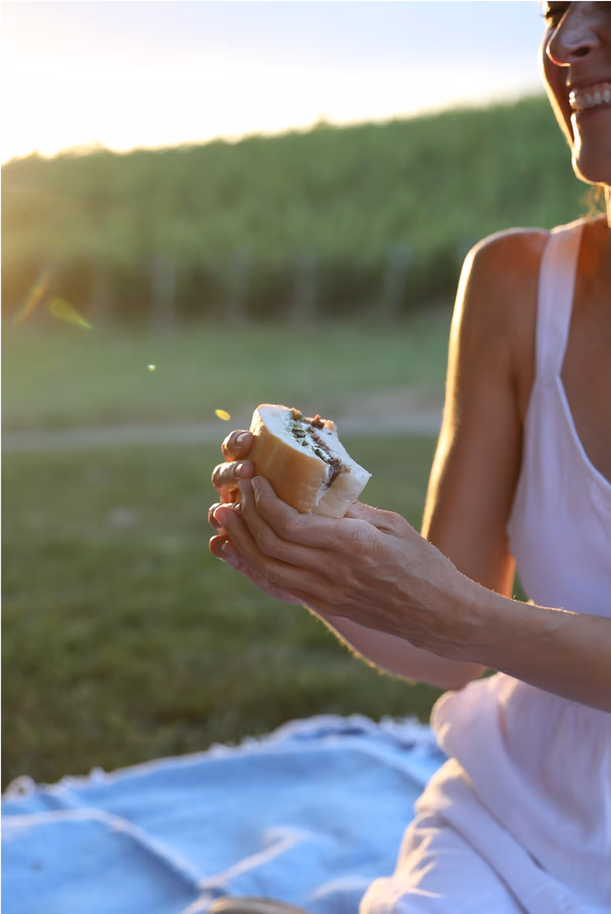 A woman in a white dress sits on a blue picnic blanket outdoors at sunset, smiling and holding a sandwich with various fillings. The background is green and sunlit.