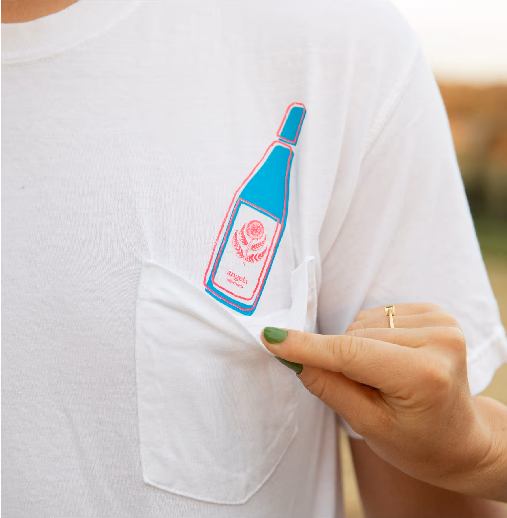 A person with green nail polish pulls open the pocket of a white t-shirt, revealing a colorful graphic of a blue and pink bottle printed above the pocket.