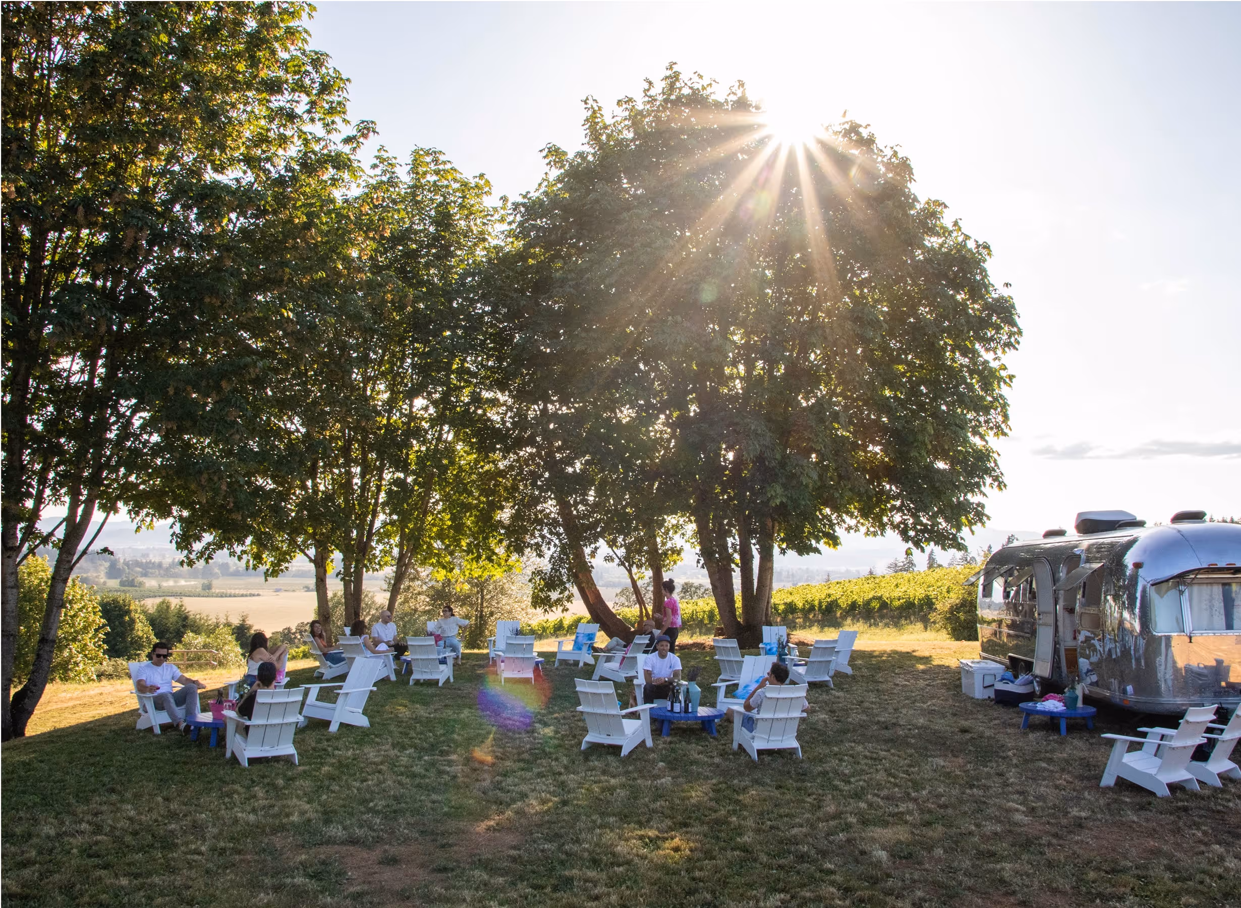 People sit on white chairs under leafy trees near an Airstream trailer, enjoying a sunny outdoor gathering with a scenic field in the background. Sunlight filters through the branches above.