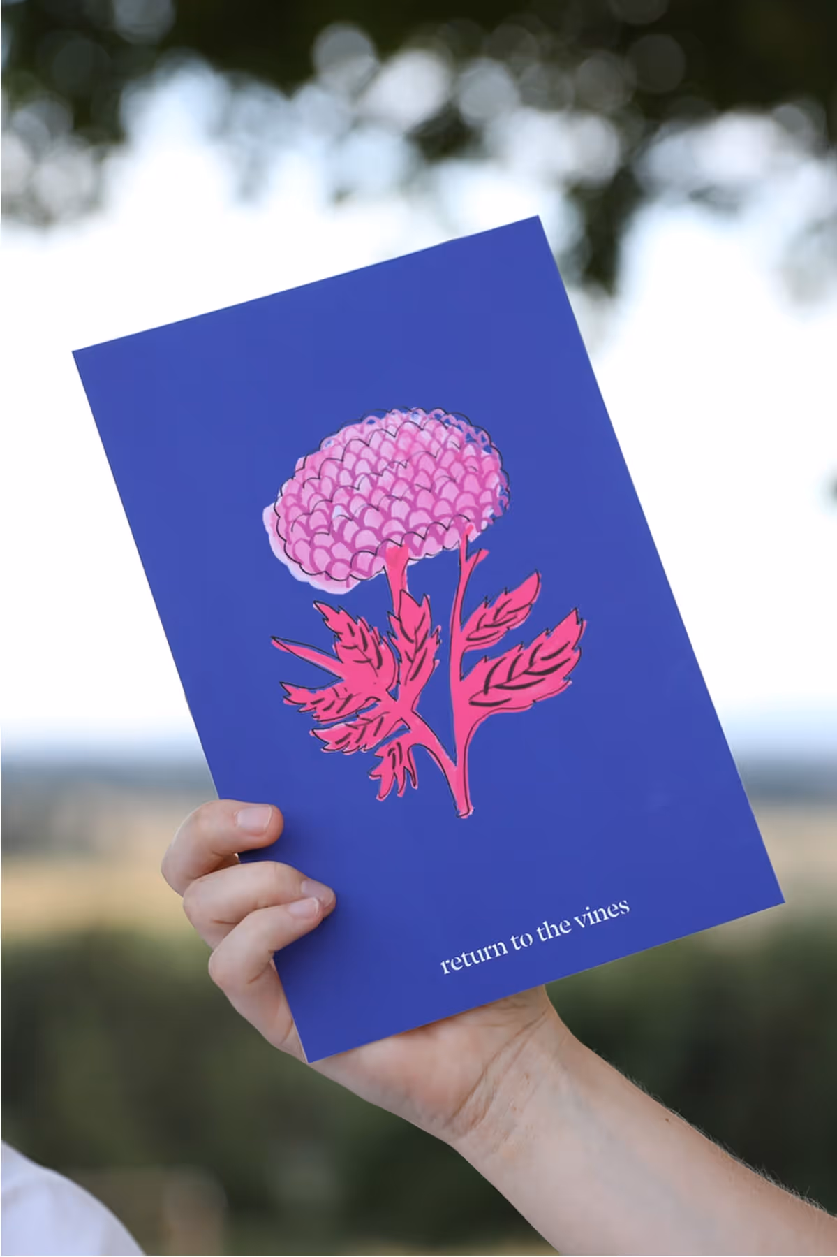 A hand holds a blue card with a pink flower illustration and red leaves. The card has the text return to the vines at the bottom. The background shows a blurred outdoor landscape.