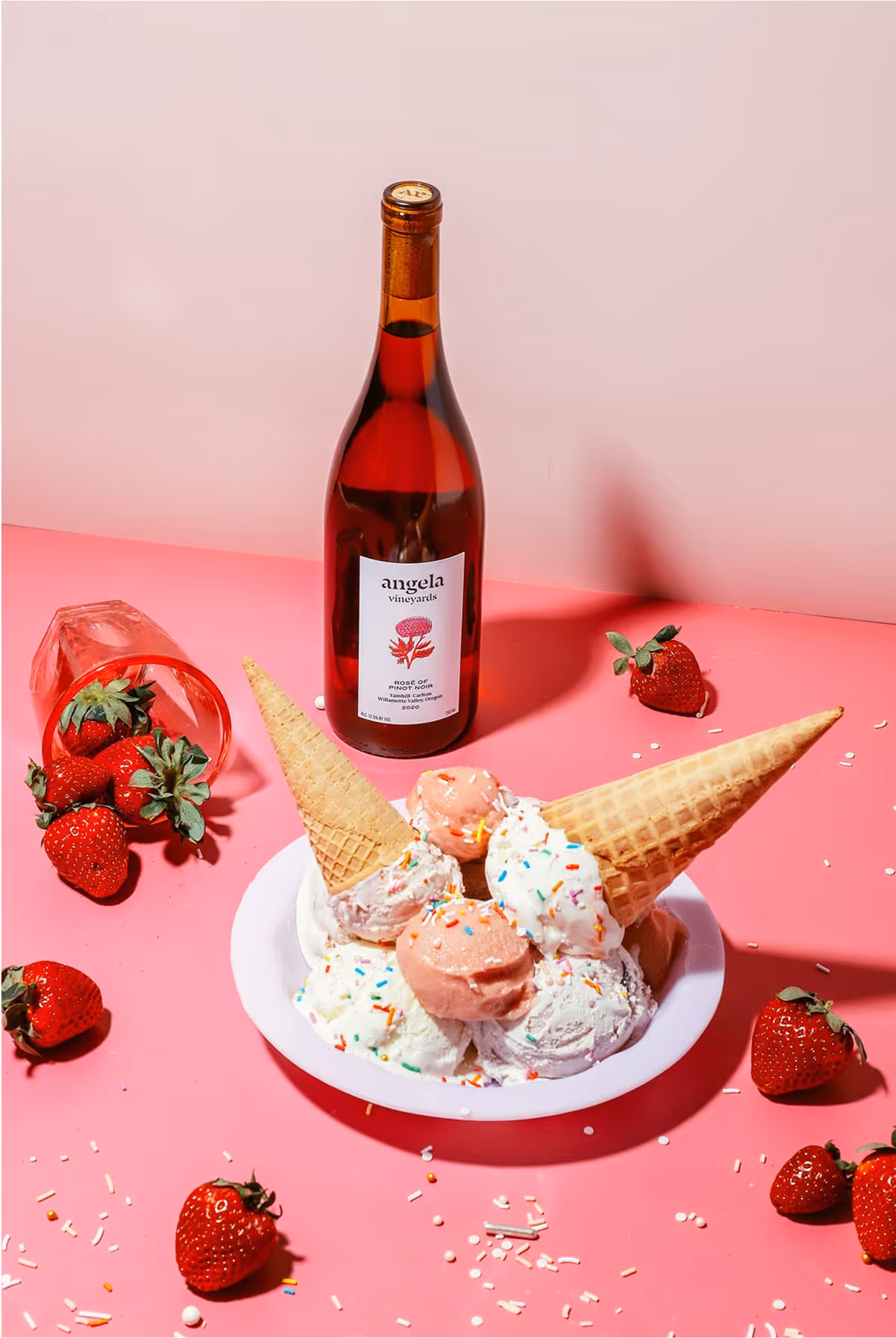 A plate of ice cream with sprinkles, topped with two waffle cones and a macaron, sits on a pink surface next to fresh strawberries, scattered sprinkles, a wine bottle, and a tipped-over wine glass.