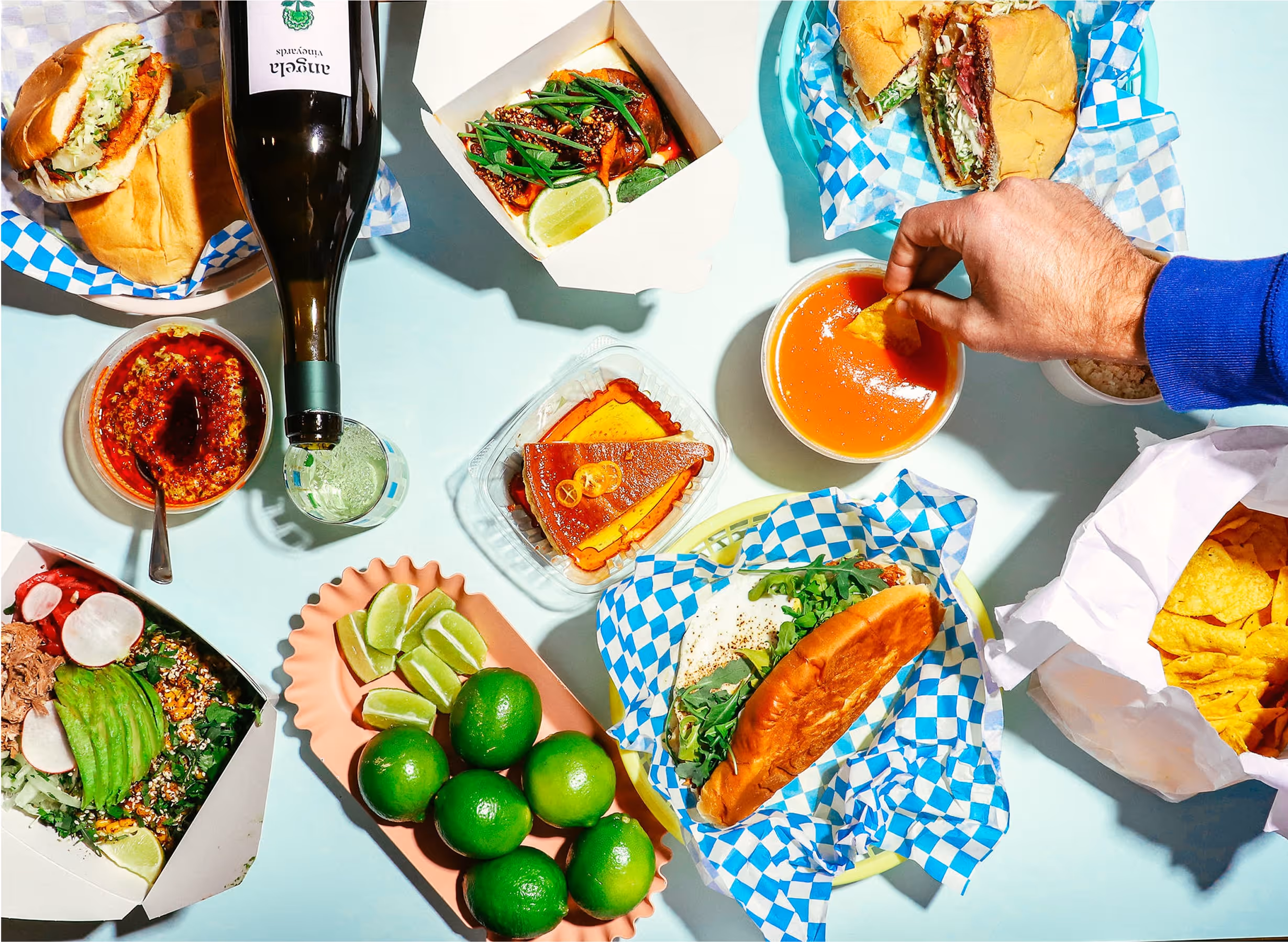 A colorful spread of takeout food, including sandwiches, tacos, chips, dip, lime wedges, a slice of cake, a bottle of wine, and a hand dipping a chip into sauce, all arranged on a light blue table.