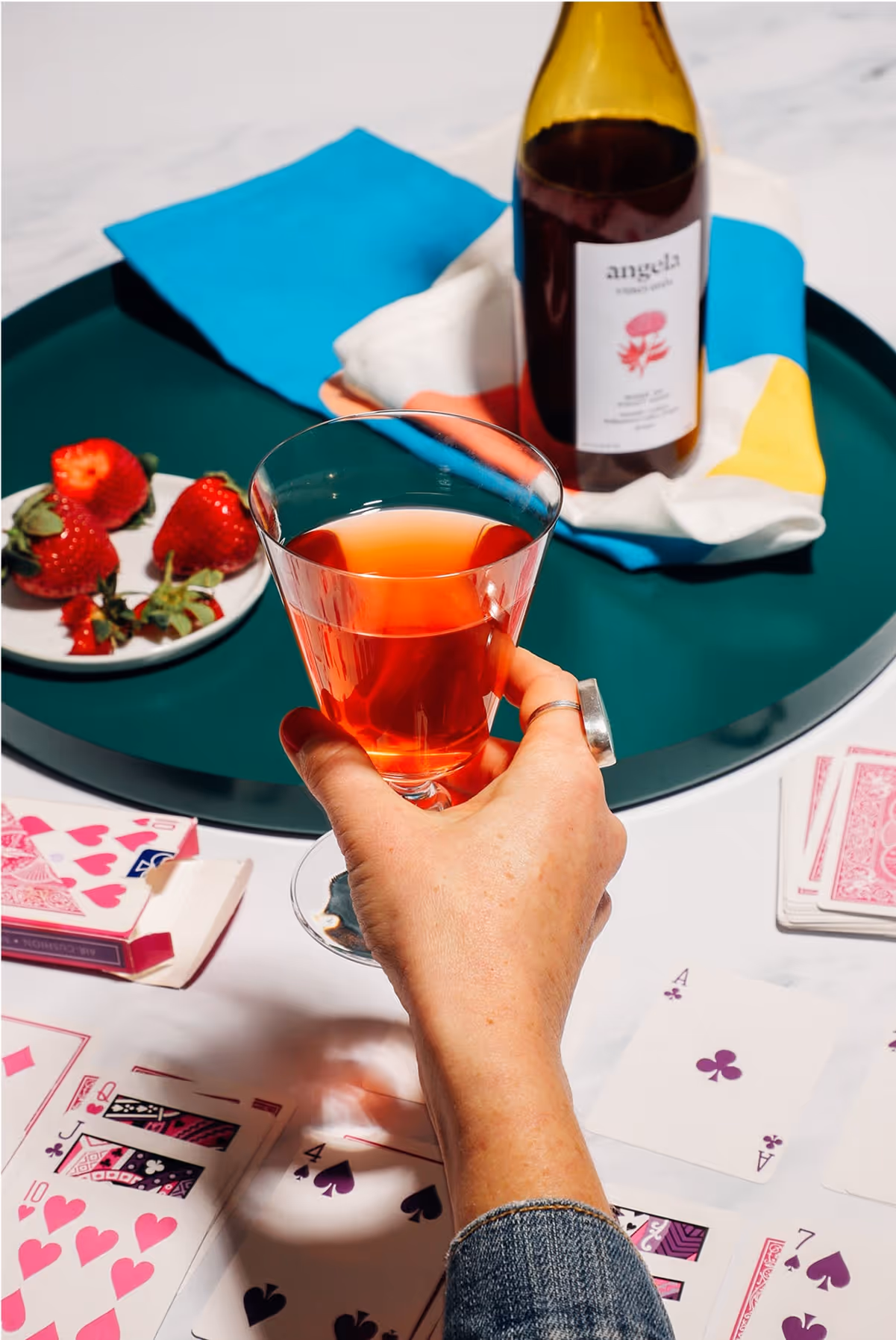 A hand holds a glass of rosé wine above a table with playing cards, strawberries on a plate, and a wine bottle on a green tray with colorful napkins.