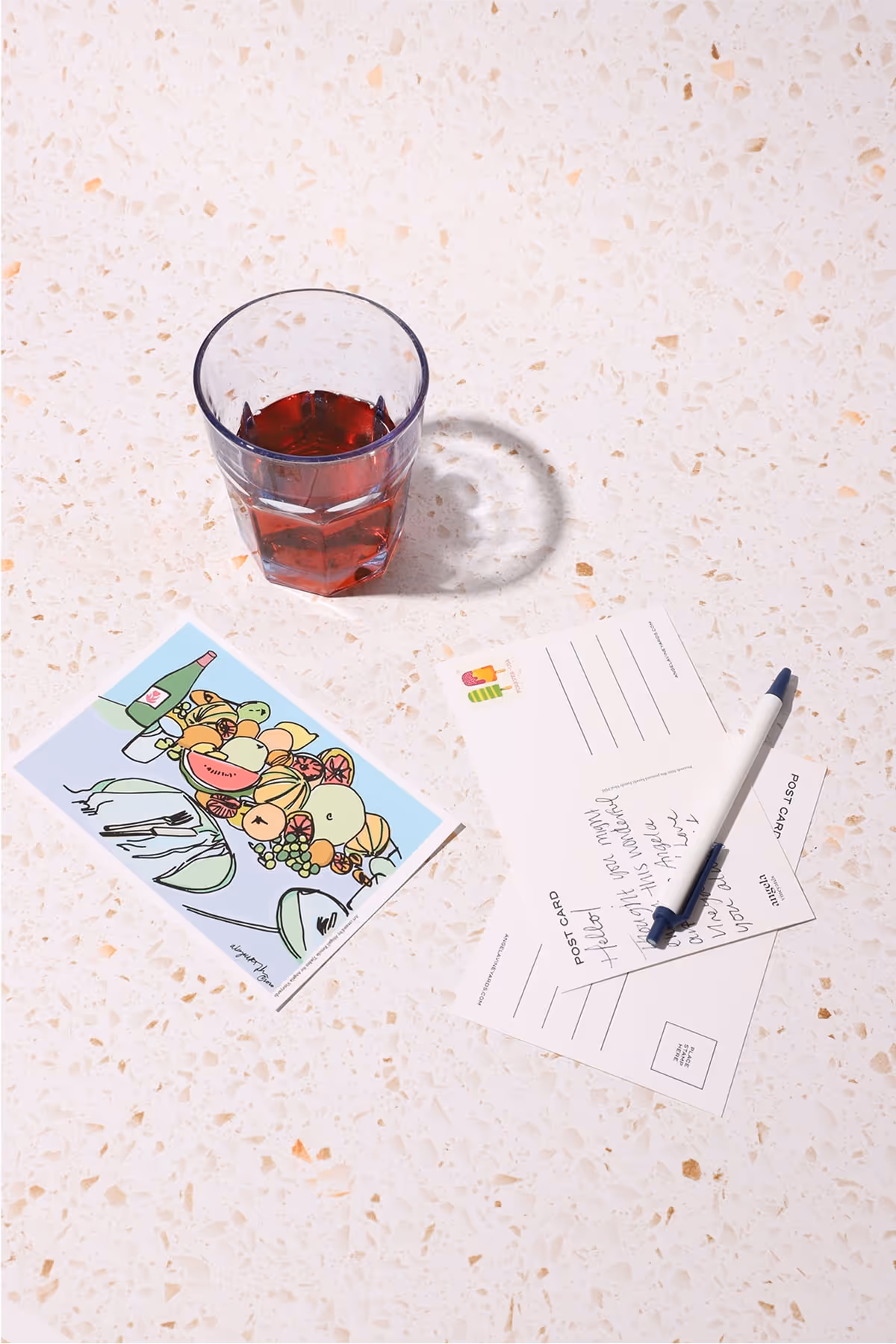 A glass of red drink sits on a terrazzo surface next to two postcards, one illustrated with cartoon animals and the other handwritten, with a pen placed on top.