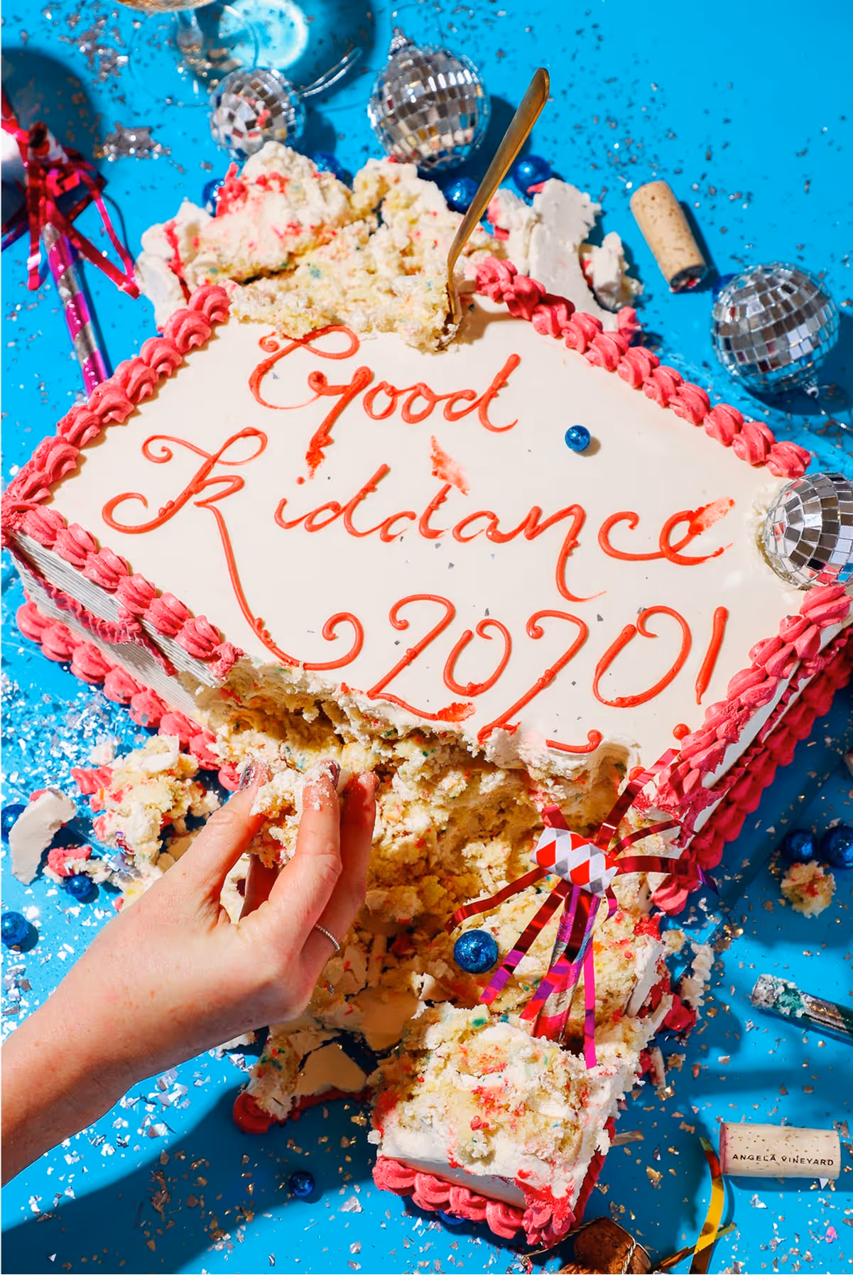 A rectangular cake with “Good Riddance 2020!” written in red icing sits on a bright blue table. The cake is partly smashed, with a hand grabbing some, surrounded by decorations, disco balls, and scattered crumbs.