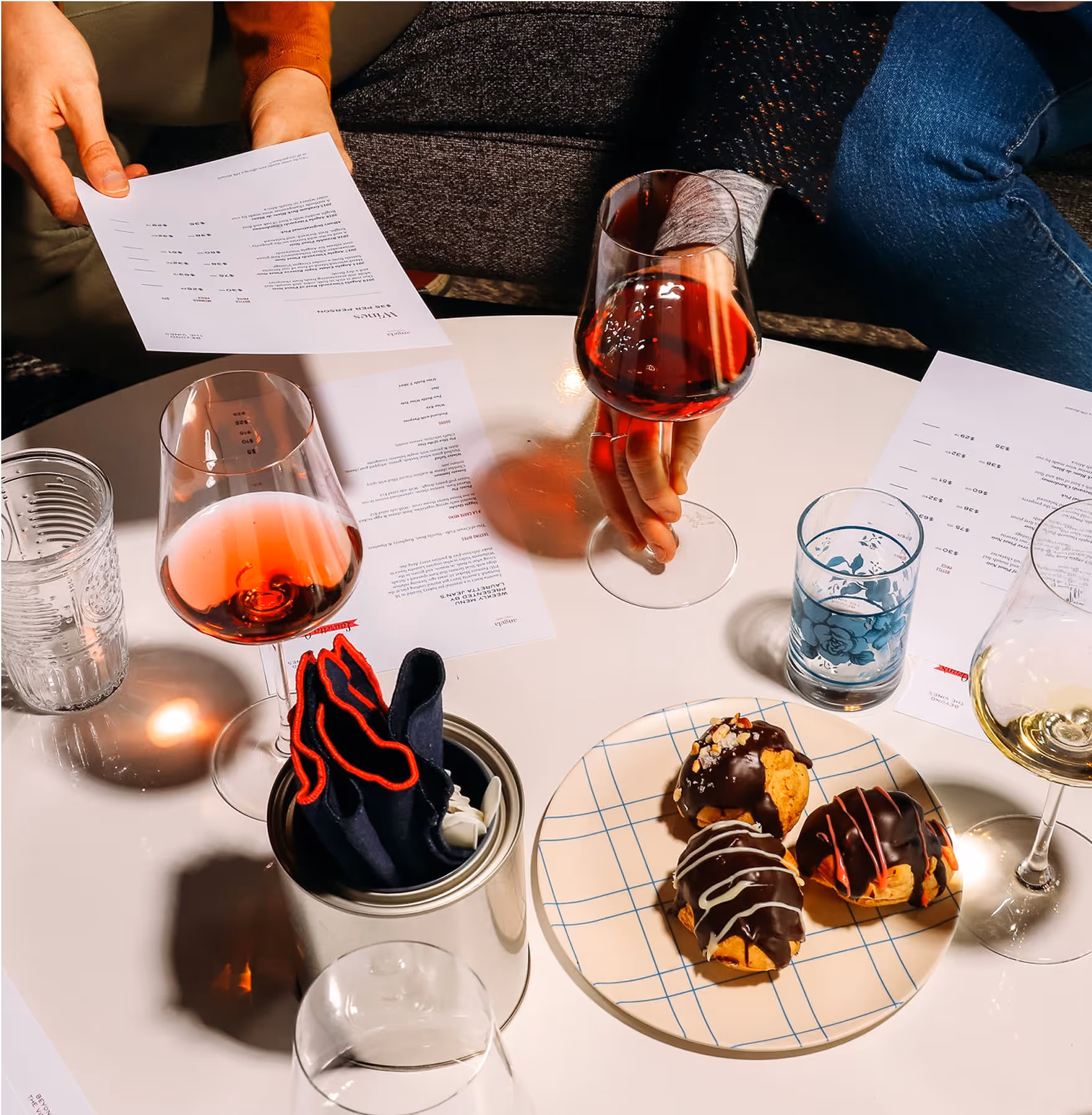 A table set with wine glasses, water glasses, menus, and a plate of chocolate-covered pastries. Two people are holding menus and wine glasses, and napkins are placed in a tin container on the table.