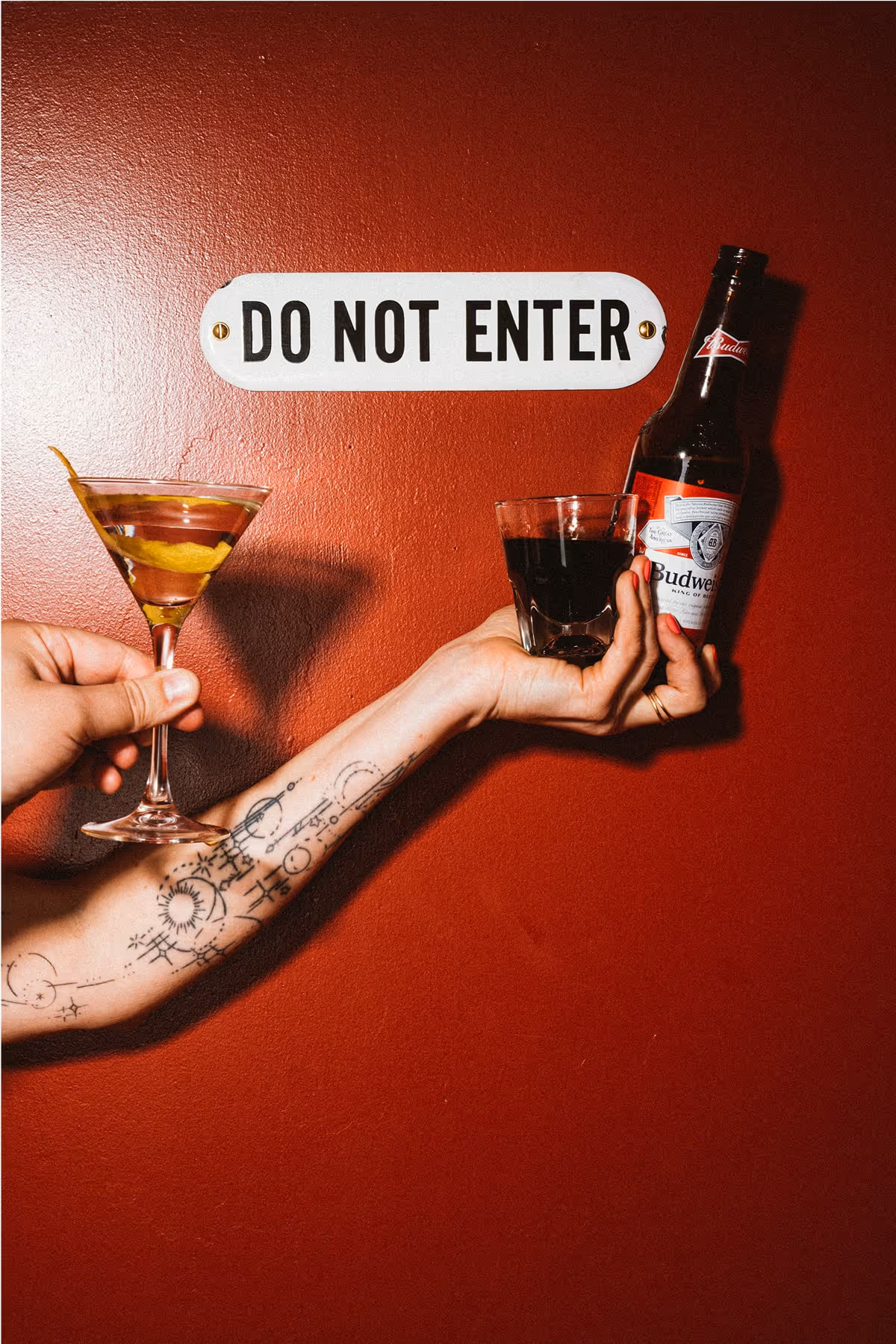 A tattooed arm holds a martini, a glass of dark liquor, and a Budweiser beer bottle against a red wall with a DO NOT ENTER sign above.