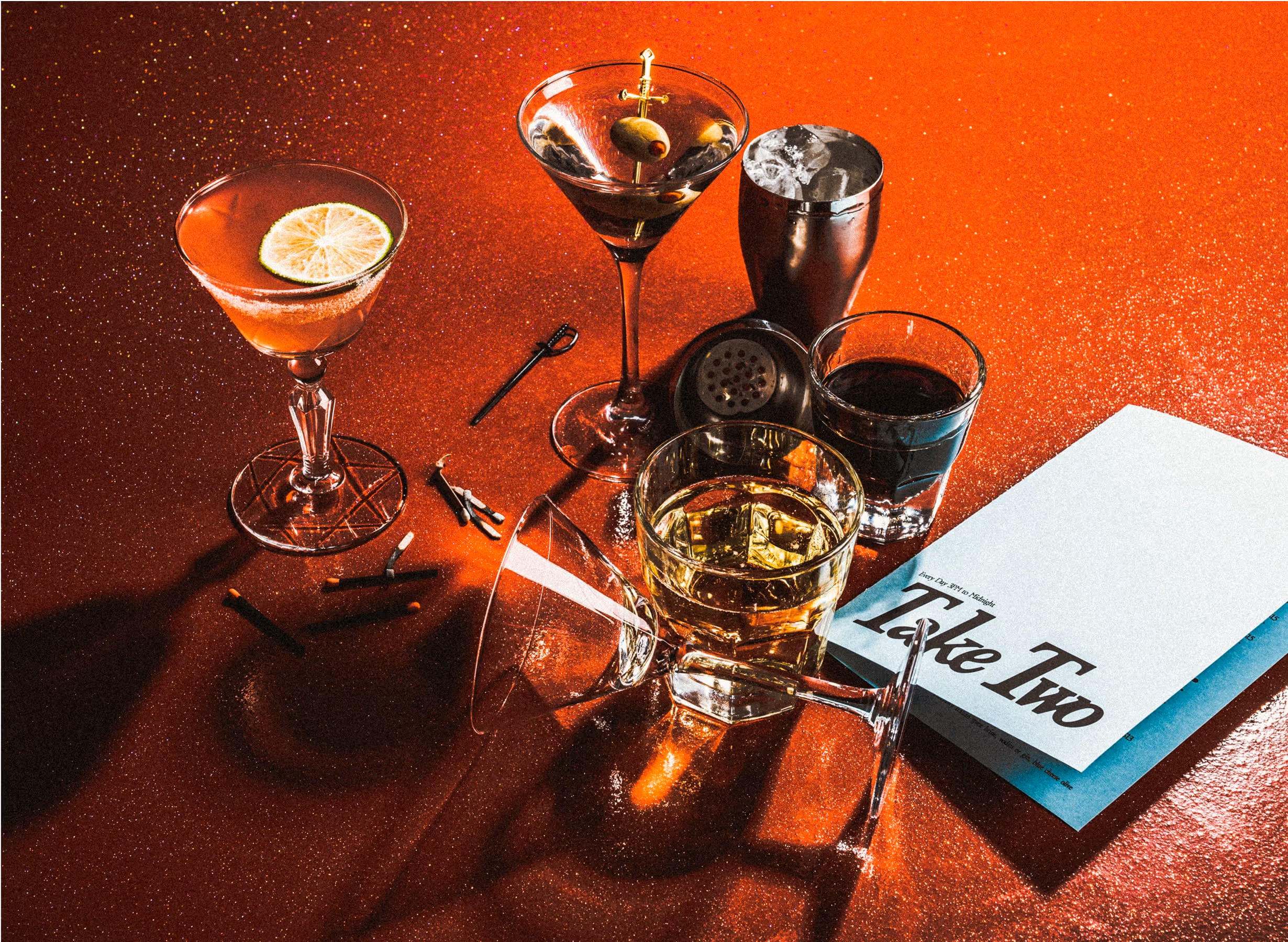 Assorted cocktails and a whiskey glass on a shimmering red surface with a metal shaker, cocktail picks, and a blue menu titled Take Two nearby, all dramatically lit.
