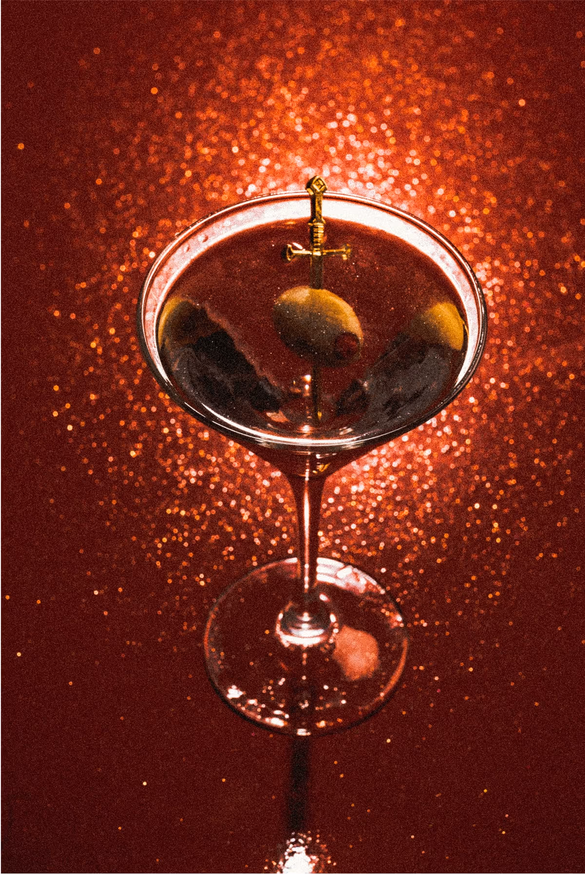 A martini glass filled with a dark cocktail, garnished with an olive and lemon twist skewered on a cocktail pick, sits on a glittery red surface with dramatic lighting.