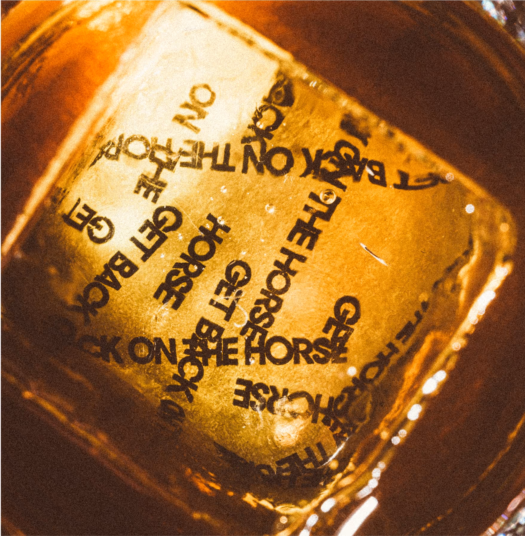 A large ice cube in a drink, viewed from above, with the phrase GET BACK ON THE HORSE repeatedly printed on its surface in black letters. The liquid around the ice cube is amber in color.