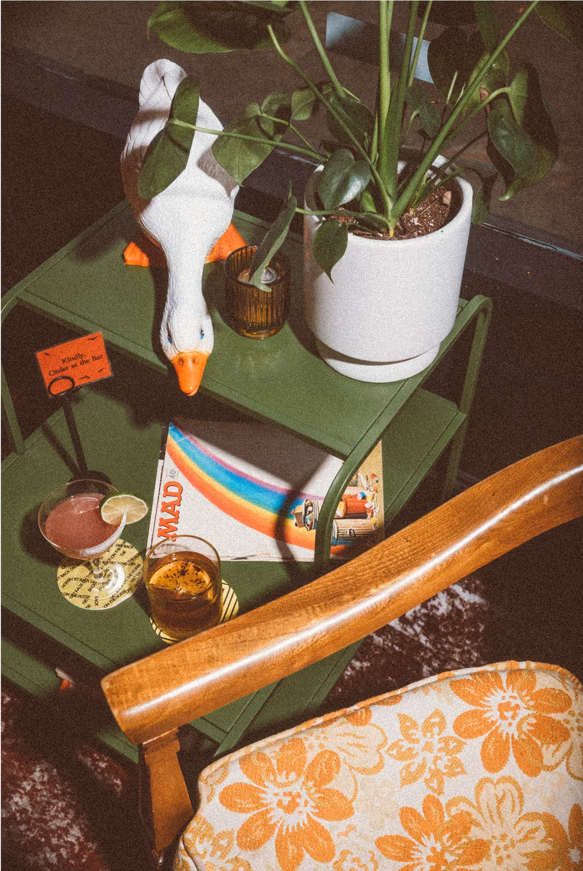 A green side table holds a potted plant, a white ceramic goose, drinks, coasters, a rainbow-covered book, and a candle. Part of a wooden armchair with orange floral upholstery is visible in front.