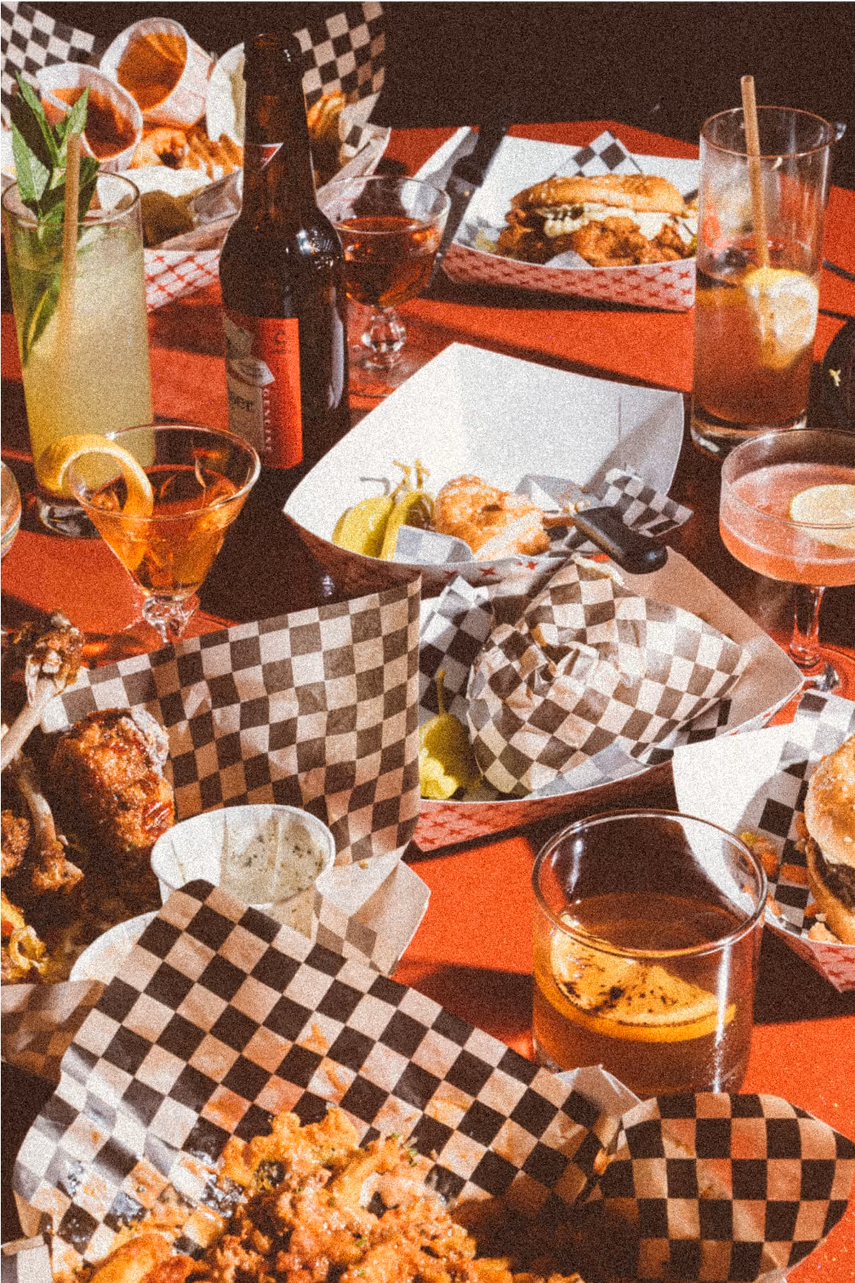 A red table covered with drinks, burgers, chicken wings, pickles, sauces, and checkered paper baskets, suggesting a lively meal with friends at a casual restaurant.