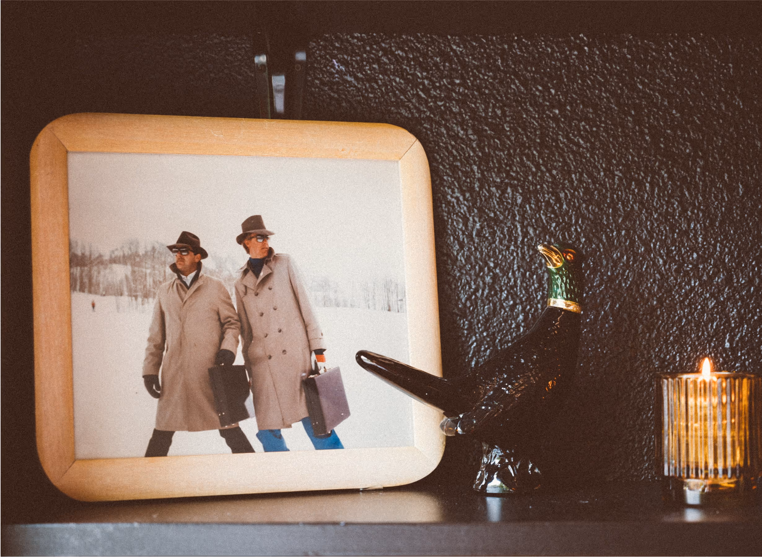A framed photo of two people in trench coats and hats walking in snow, holding suitcases. Next to the frame are a black bird figurine and a lit candle in a glass holder on a dark shelf.