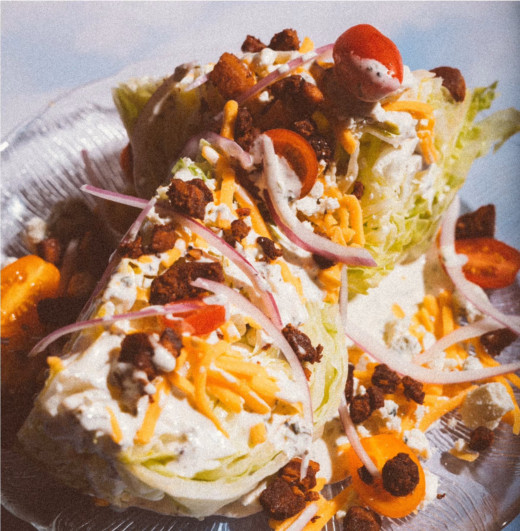 Two wedge salads with lettuce, cherry tomatoes, shredded cheese, red onion slices, blue cheese crumbles, bacon bits, and creamy dressing, served on a silver platter.