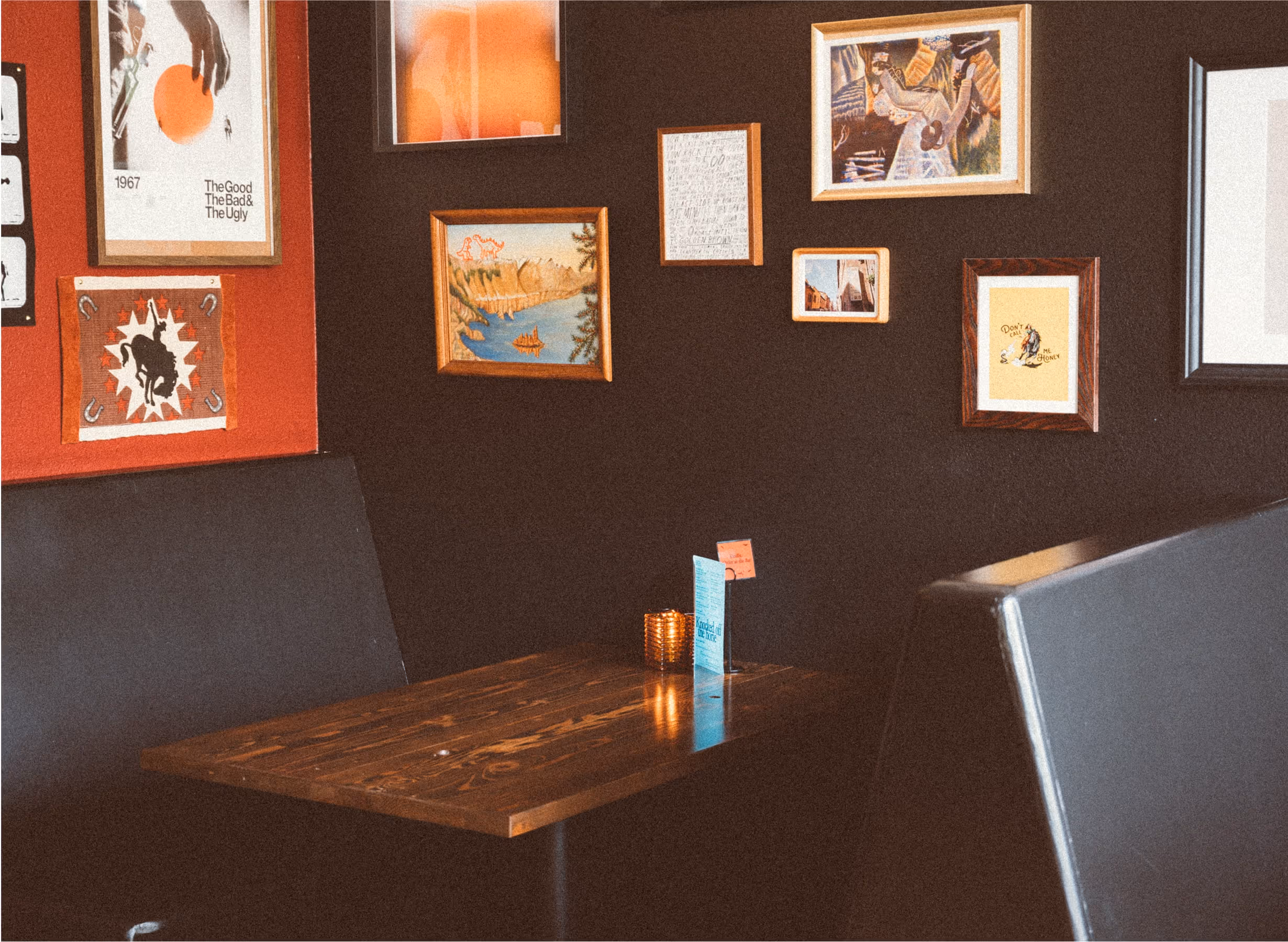 A cozy restaurant booth with a dark wooden table, black padded seats, a teal menu, a candle holder, and various colorful framed artworks on a black and red wall.