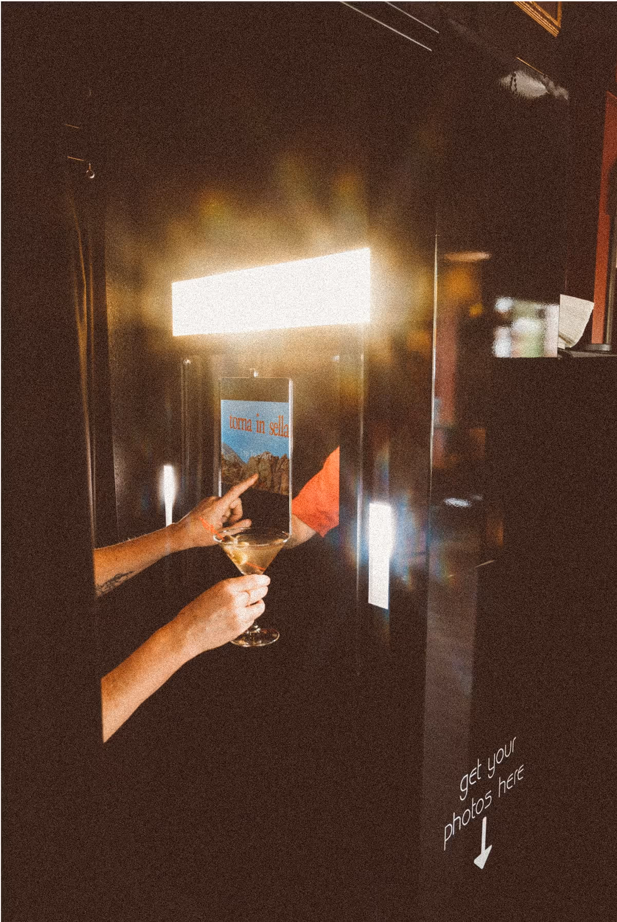 A person holds a wine glass and reaches toward a photo booth slot, with a glowing light above and a sign reading get your photos here and an arrow pointing down.