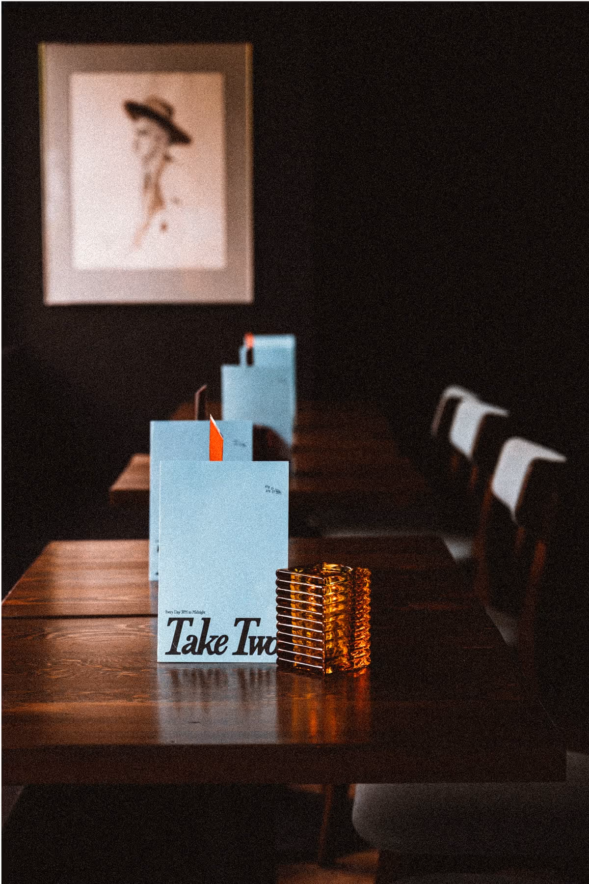 A dimly lit restaurant with wooden tables, blue menus, a candle holder, and chairs. A framed portrait of a woman in a hat hangs on the dark wall in the background.