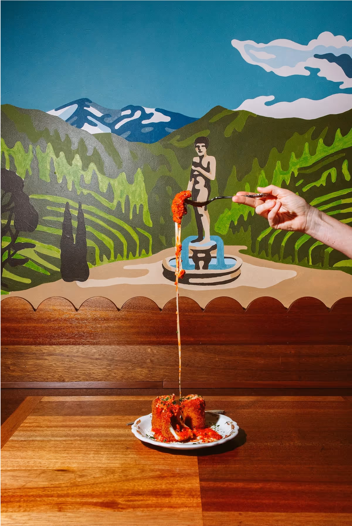 A hand lifts a forkful of cheesy, saucy lasagna high above a plate on a wooden table. Behind, a colorful mural shows green hills, a statue, and mountains under a blue sky.