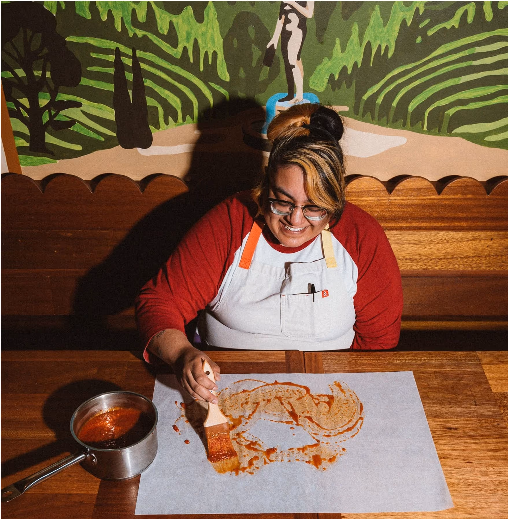 A person wearing glasses and a red-and-white shirt smiles while spreading sauce on parchment paper with a brush, sitting at a wooden table with a saucepan of sauce nearby. A colorful mural is in the background.