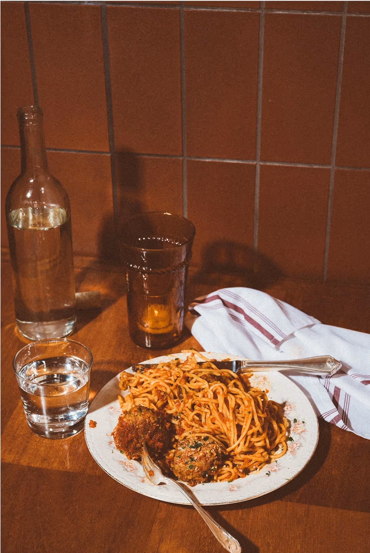 A plate of spaghetti with meatballs sits on a wooden table next to a fork, a glass of water, a clear glass bottle, an amber glass, and a white napkin with red stripes, with brown tiled wall in the background.